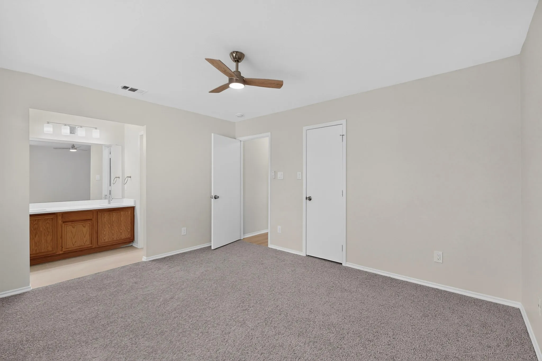 Wall-to-wall carpet flooring, ceiling fan with integrated lighting, multiple white interior doors, and a bathroom vanity with a wood-finish cabinet, dual sinks, and an overhead light fixture