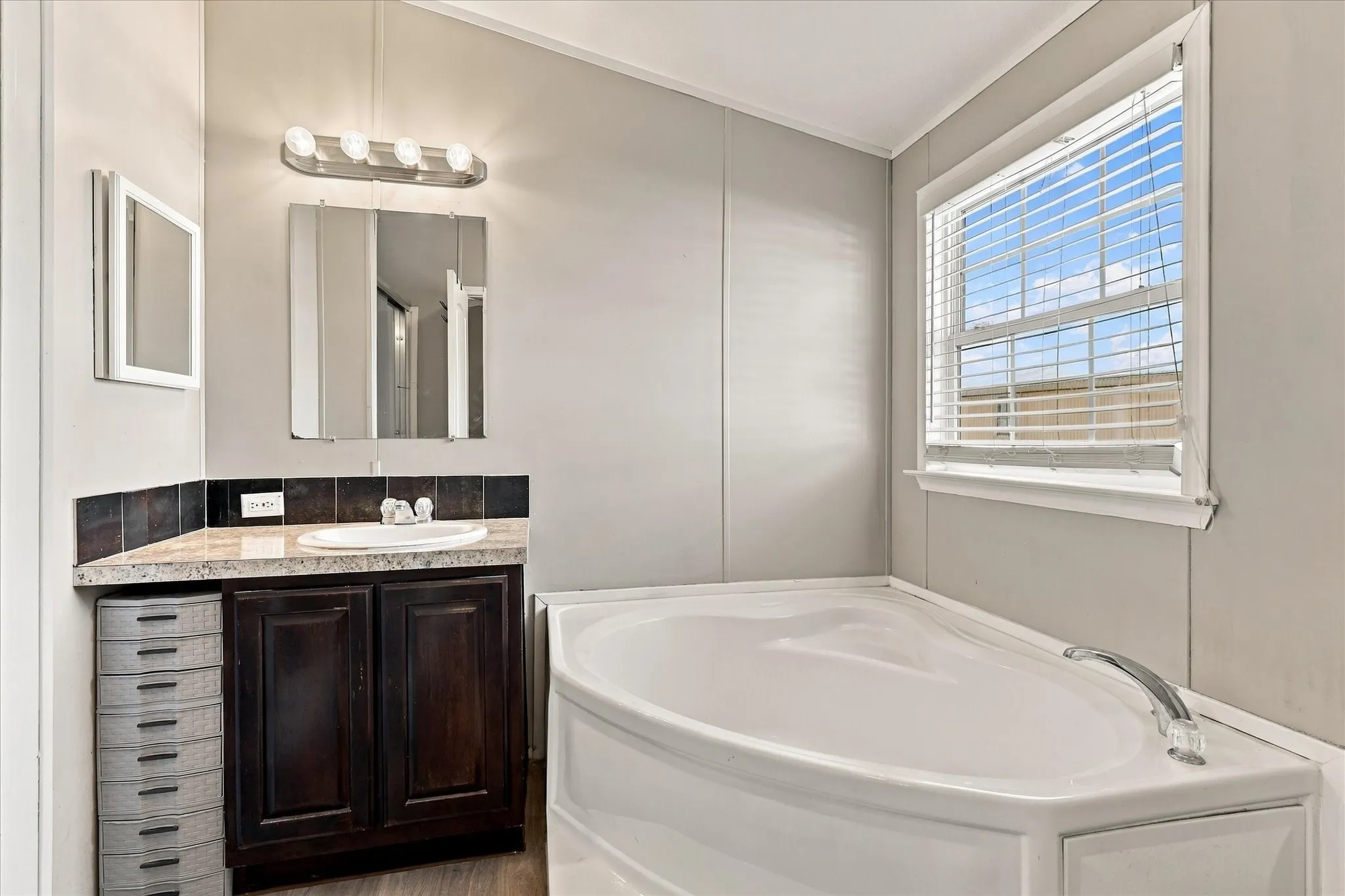 Corner soaking tub, dual medicine cabinets, vanity with countertop, dark wood-finish cabinetry, and a window with blinds