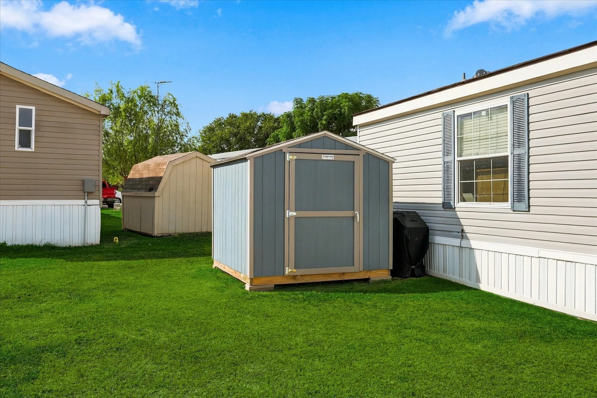 Manicured lawn with two storage sheds and a mobile home featuring siding and a window with shutters