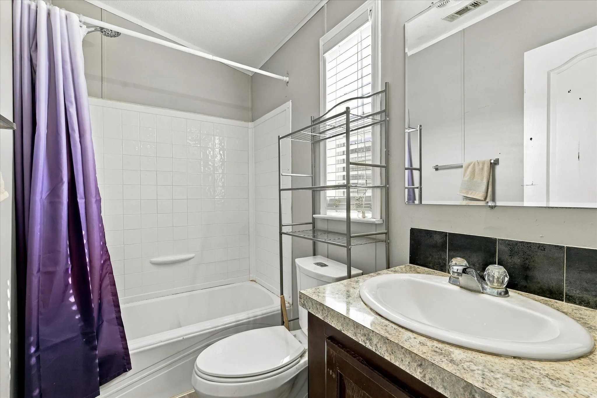 Bathroom featuring a white tub and shower combination with tile surround, a single vanity with an oval basin, a light-toned countertop, and a window with blinds