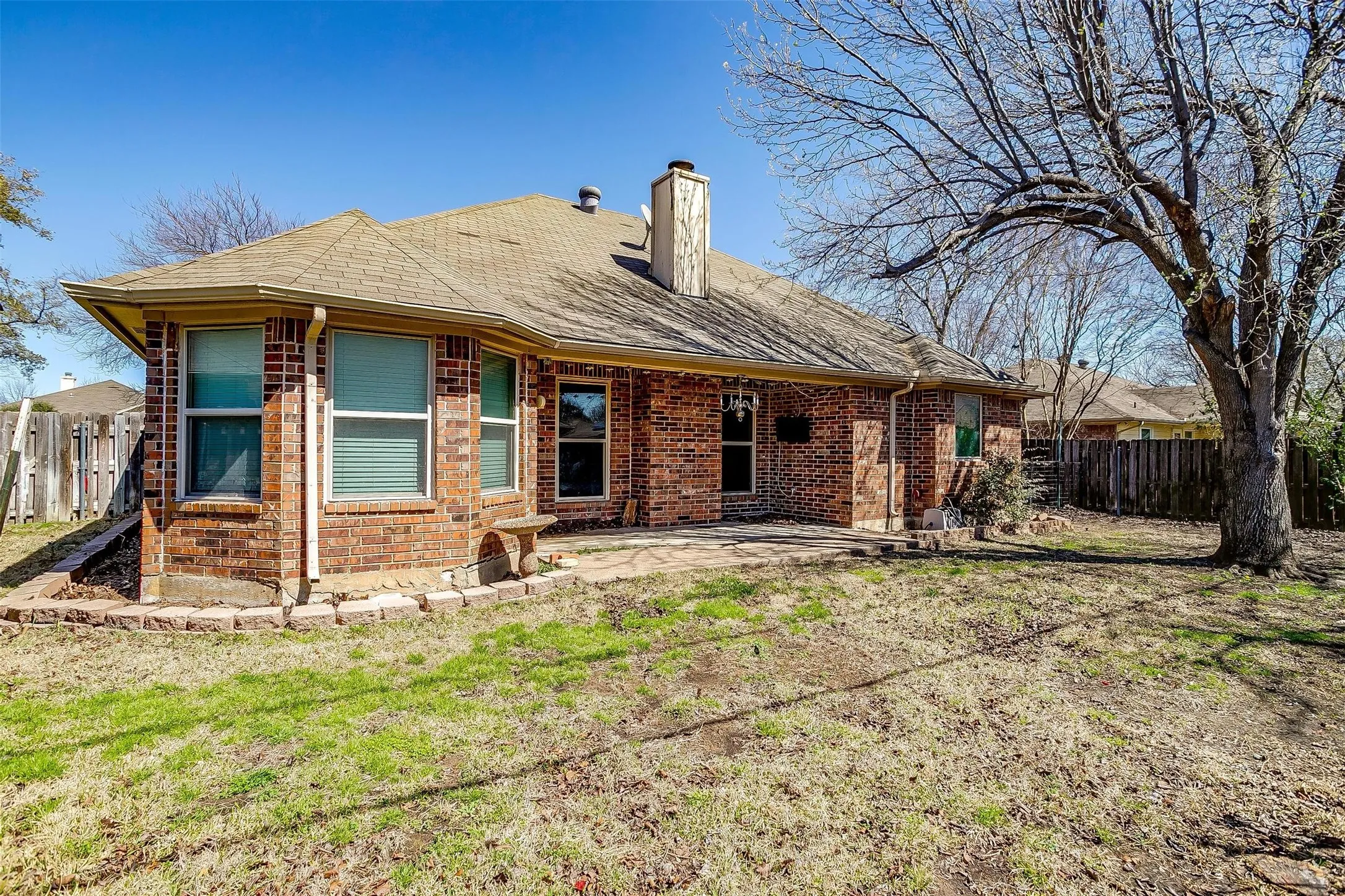 Brick exterior with a covered patio, a stone bench, multiple windows, and a large tree