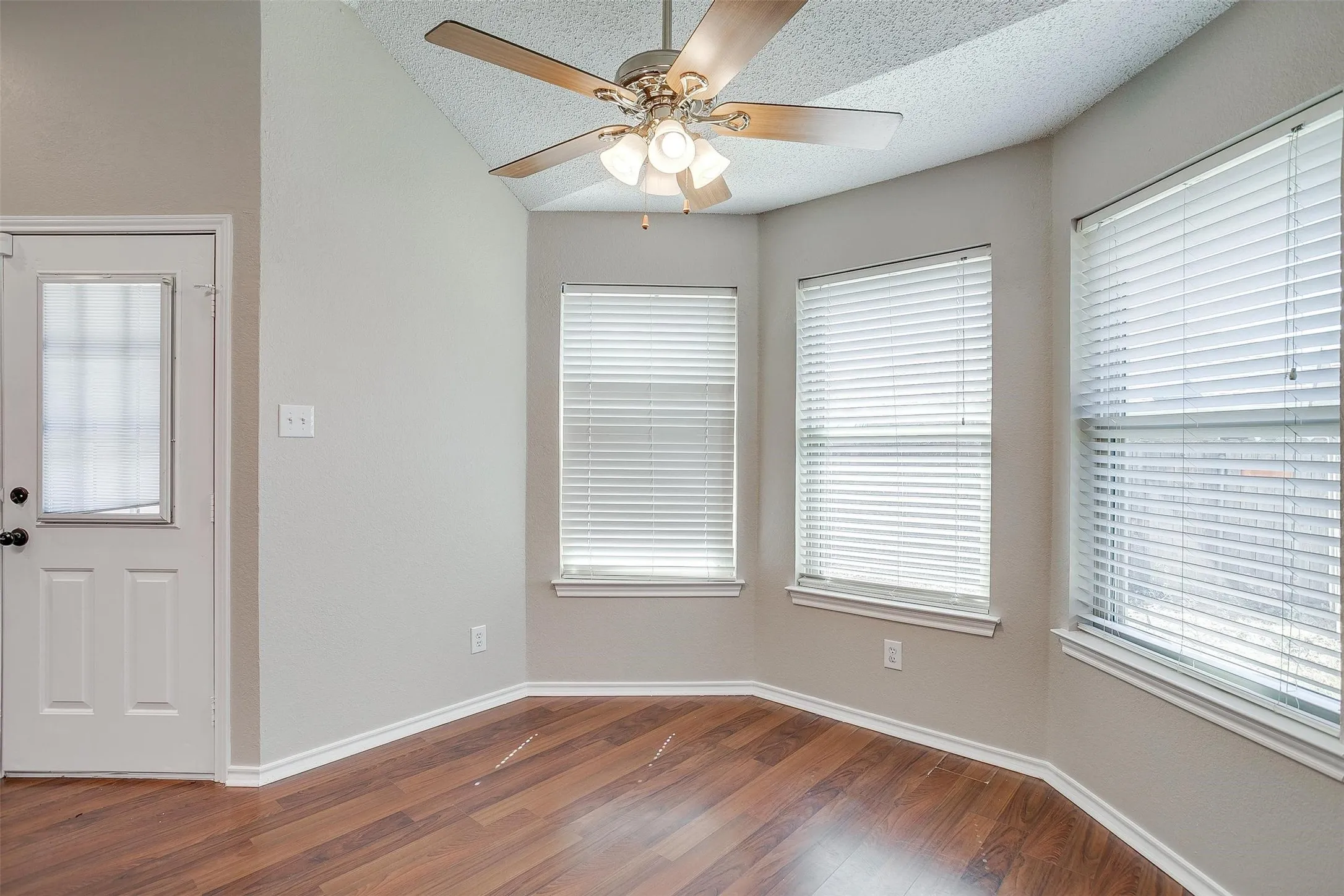 Wood-finish flooring, multiple windows with blinds, angled ceiling, ceiling fan with light fixture, and a white paneled door with glass insert