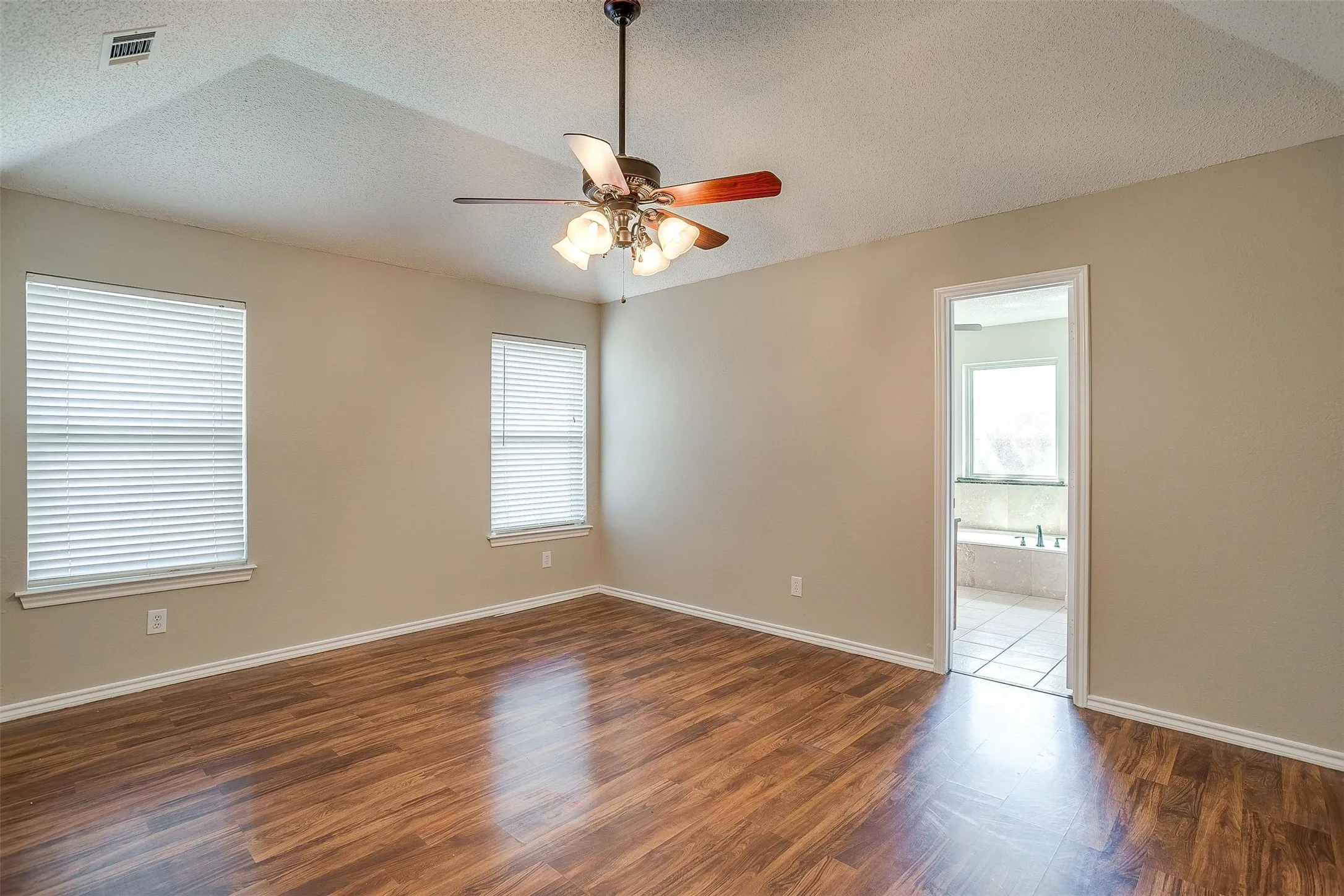 Tray ceiling with ceiling fan and light fixture, two windows with blinds, wood-finish flooring, and neutral wall tones
