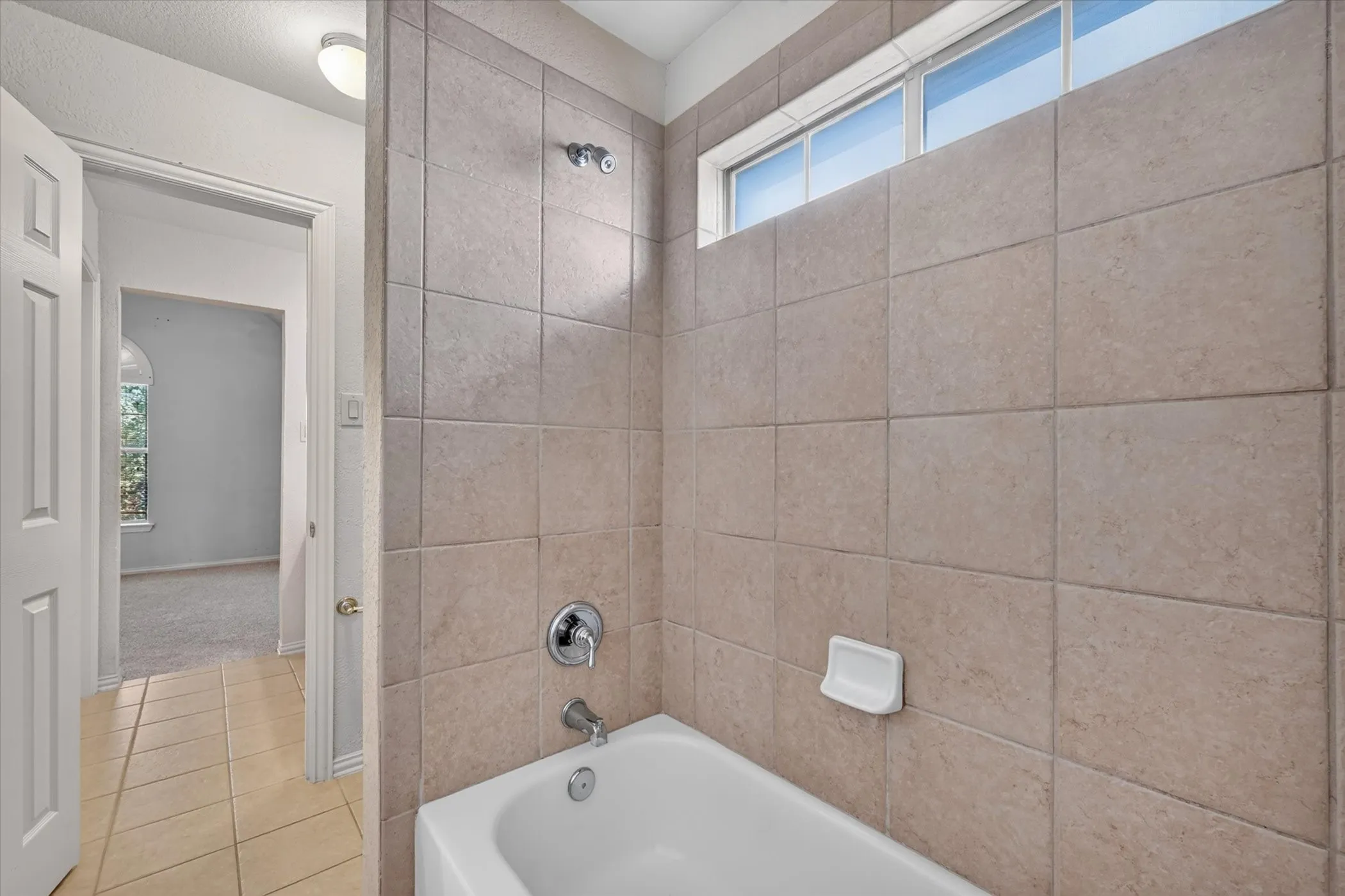 Bathtub with chrome fixtures, full-height tile surround, and an upper transom window