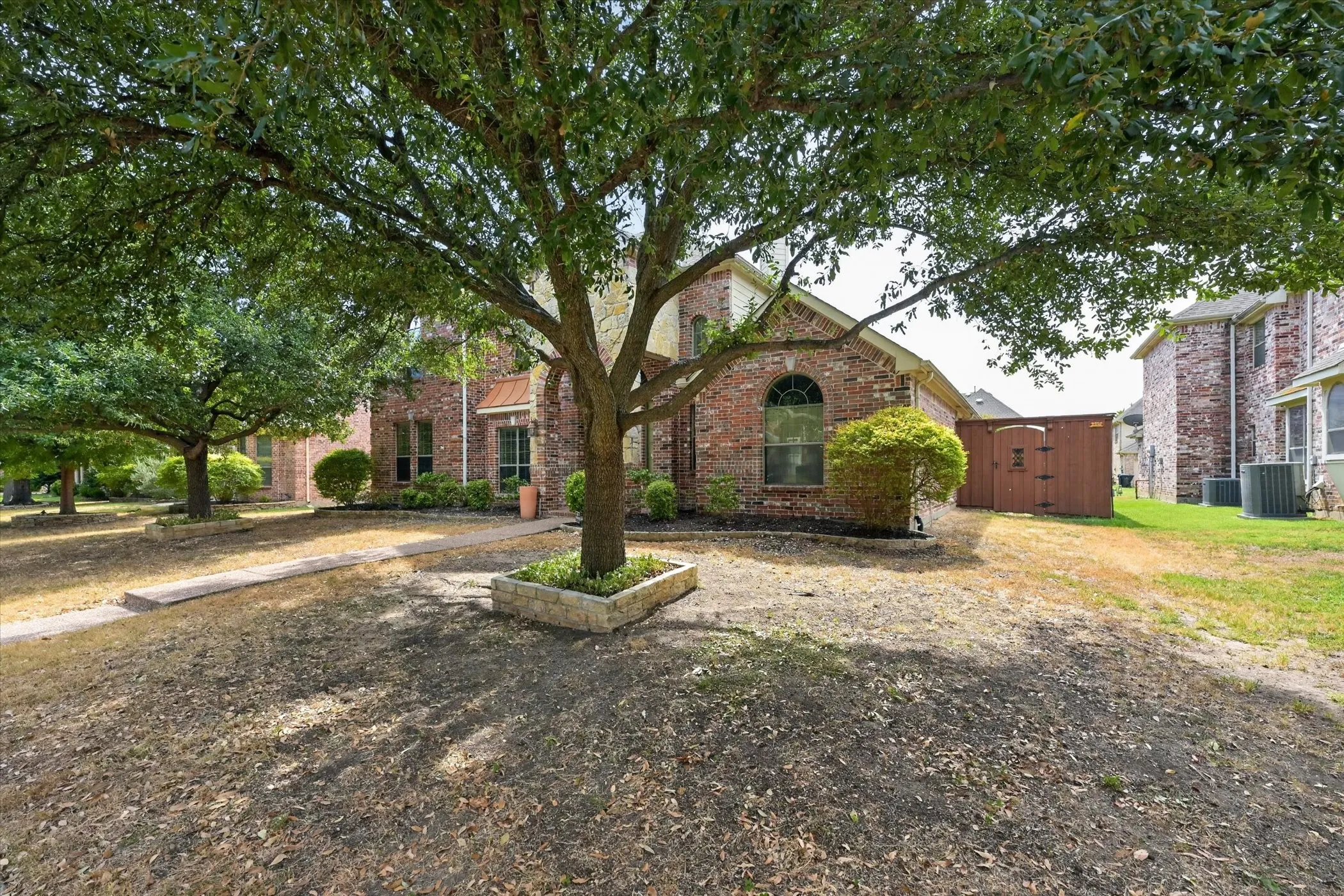 Mature trees providing ample shade, brick exterior, arched window details, stone-edged planter, and a detached storage shed
