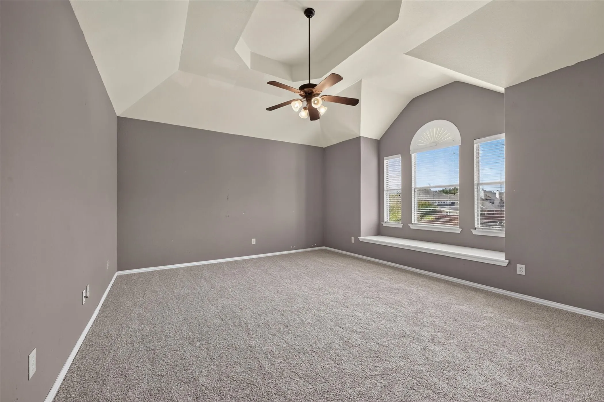 Vaulted tray ceiling with a ceiling fan, arched window, two rectangular windows, and a built-in window seat