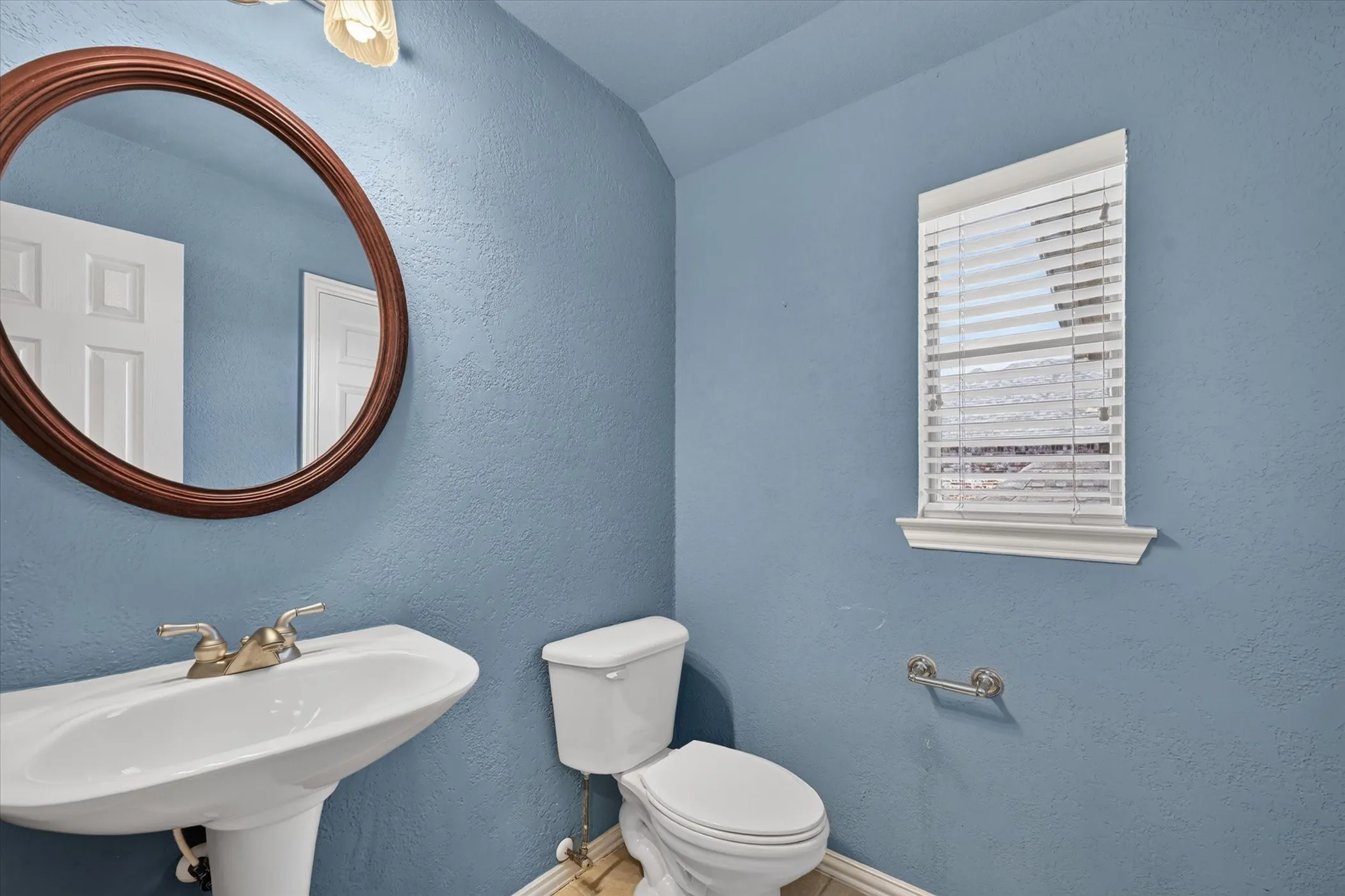 Pedestal sink with brass-finish faucet, oval wood-frame mirror, and white window blinds