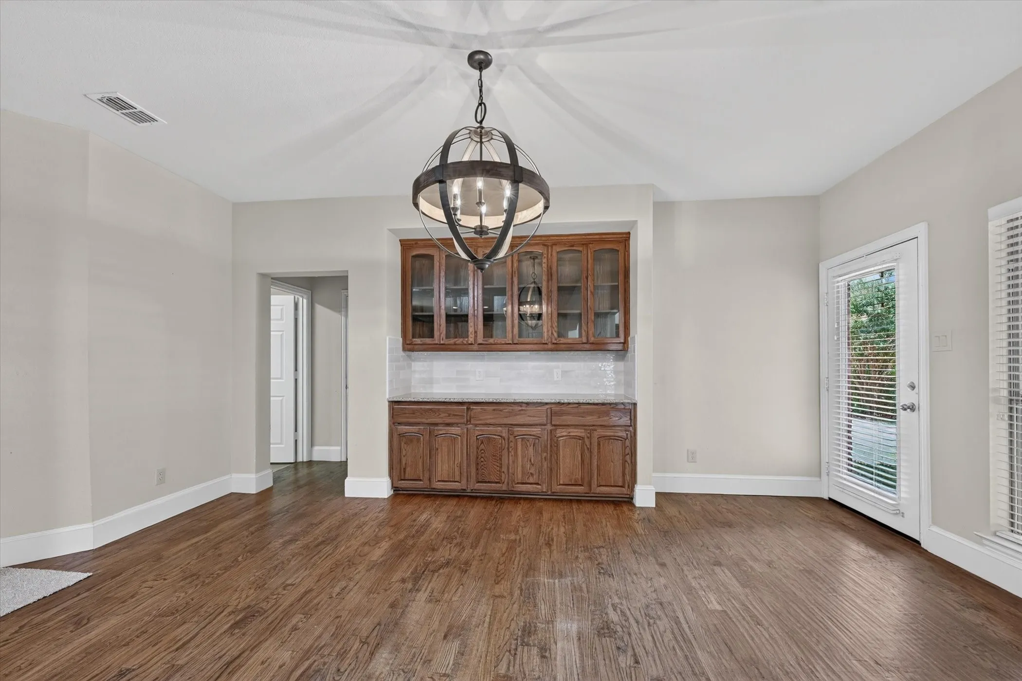 Wood-finish flooring, built-in cabinetry with glass doors, large orb chandelier, tiled backsplash, and private exterior door