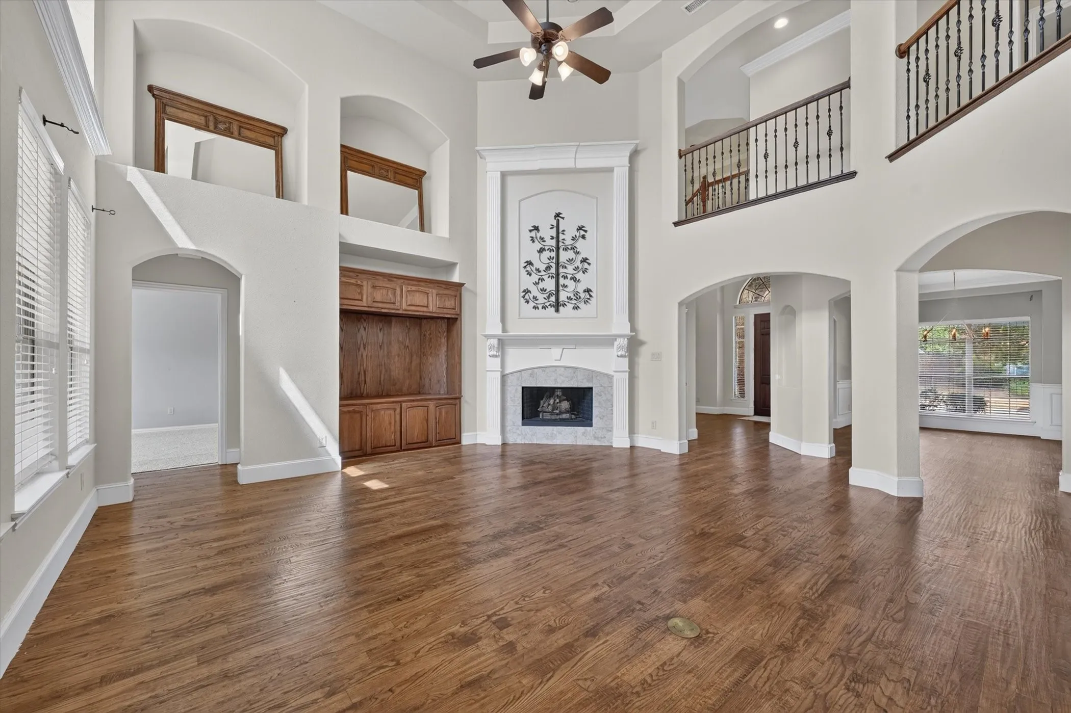 High ceilings with a ceiling fan, wood-finish flooring, white fireplace with a tiled surround, built-in wooden cabinetry, and an upper-level balcony with metal balusters