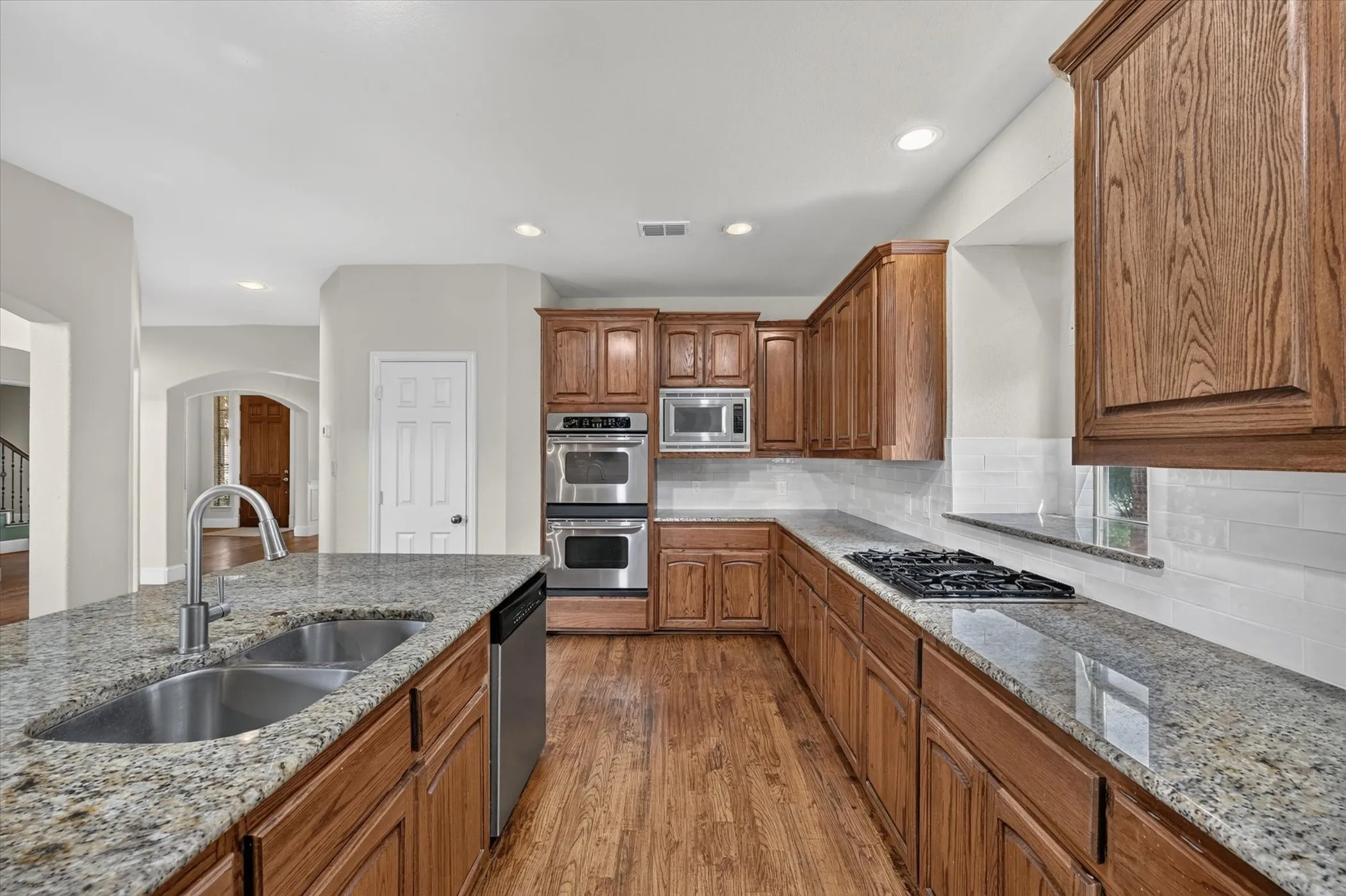 Kitchen featuring wood-finish flooring, abundant wood cabinetry, granite countertops, a double basin stainless steel sink, and stainless steel appliances