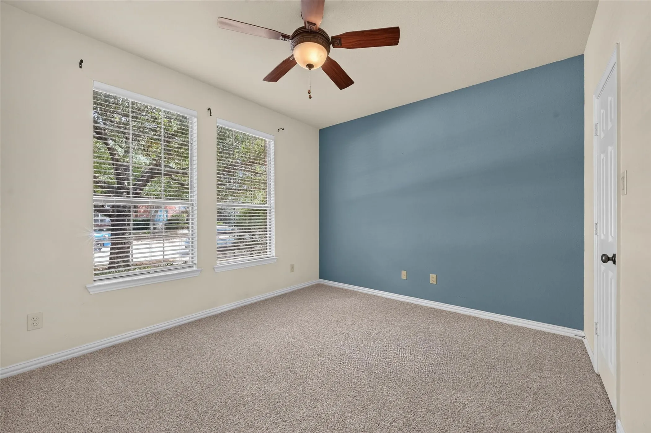Plush carpeted flooring, two windows with blinds, a wood-finish ceiling fan with integrated lighting, and a feature wall