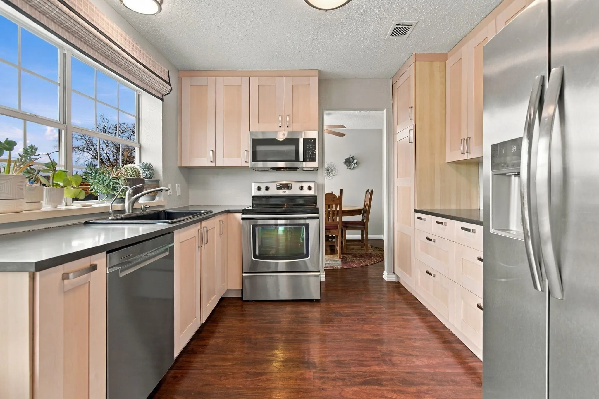 Stainless steel appliances, light wood-finish cabinetry, dark countertops, dark wood-finish flooring, and a large window