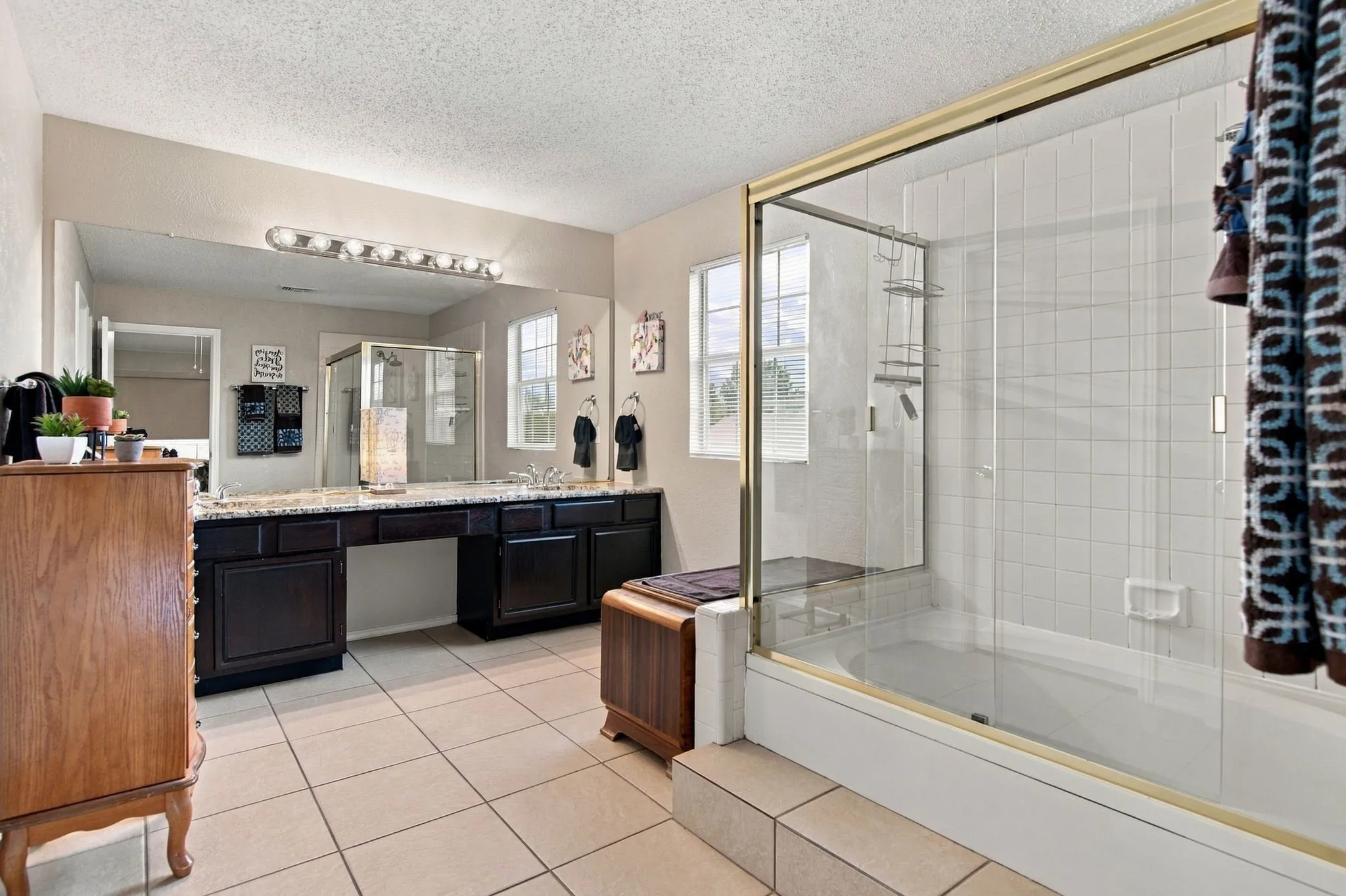 Tile flooring, dual-sink vanity with dark wood-finish cabinetry, built-in makeup vanity, large wall-mounted mirror with overhead lighting, and a bathtub with a glass enclosure