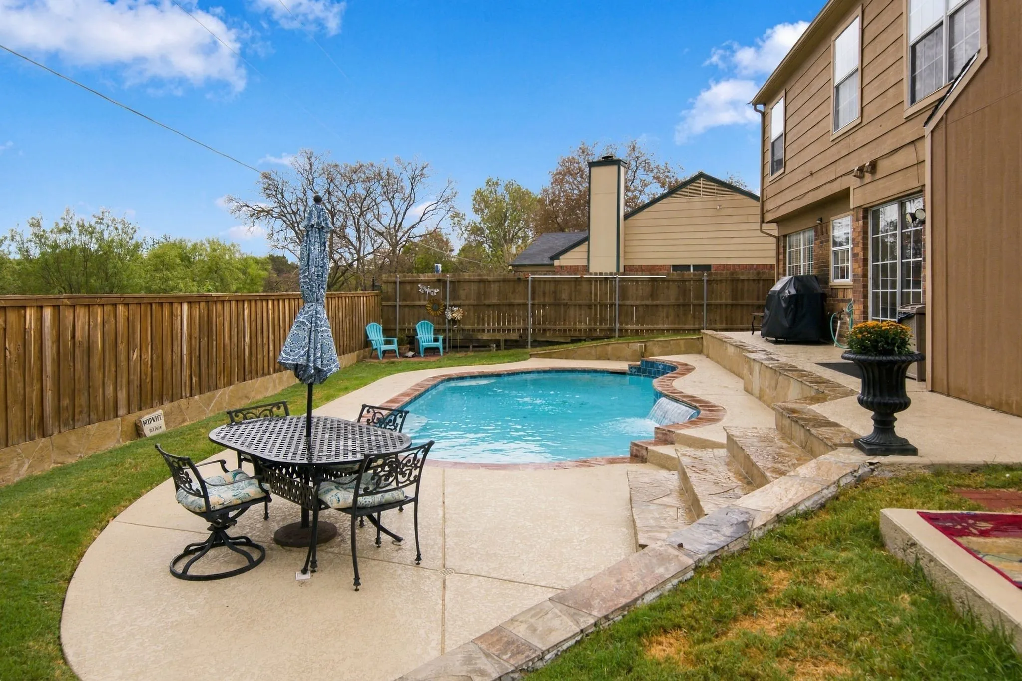 Outdoor pool with a waterfall feature, a multi-level concrete patio, retaining wall, and a privacy fence