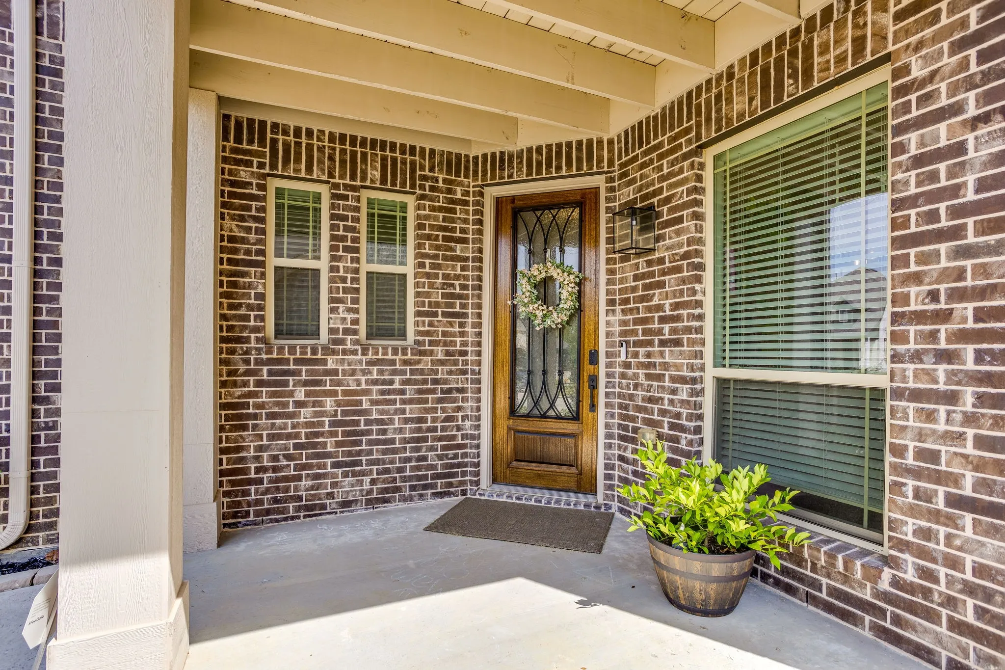 Brick exterior with white mortar, wood-paneled door with wrought iron detailing, wall-mounted exterior light fixture, and covered porch
