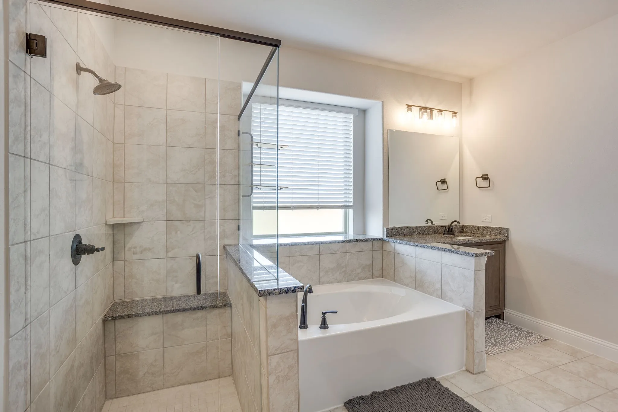 Tiled shower with glass enclosure and built-in bench, soaking tub with dark bronze fixtures, single vanity with countertop, and window with blinds