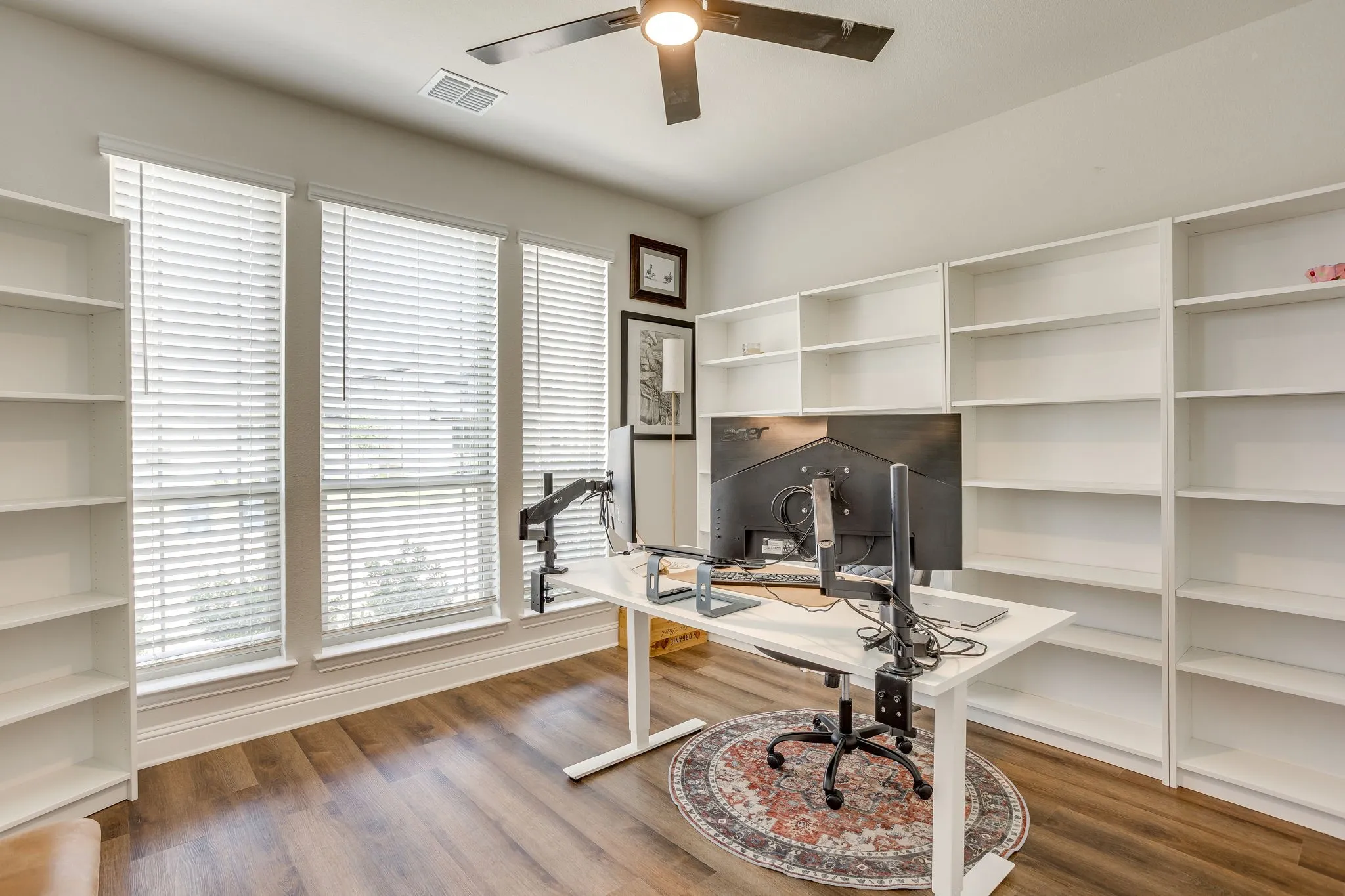Wood-finish flooring with white baseboards, three windows with blinds, a ceiling fan, and extensive built-in white shelving