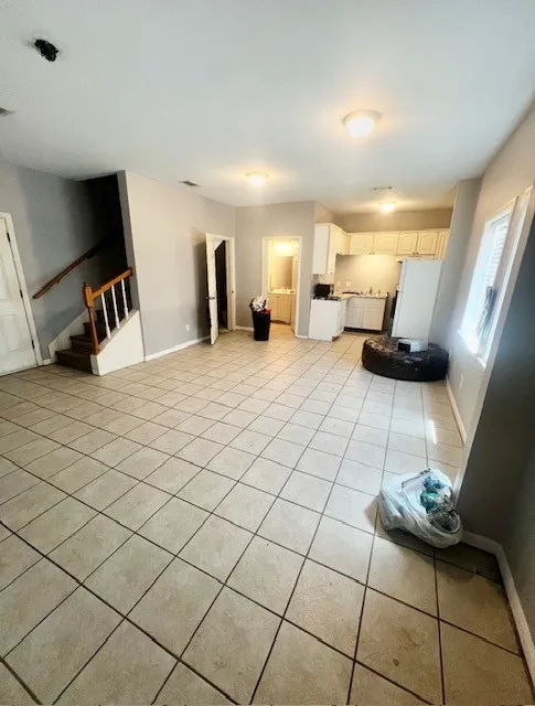 Expansive main living area with neutral-toned tile flooring throughout, featuring a staircase with wood railings, a kitchen with white cabinetry, and a bathroom with a vanity and mirror