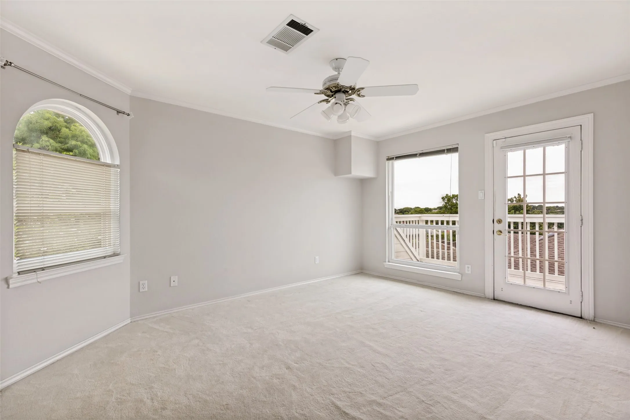 Carpeted room featuring a ceiling fan, crown molding, an arched window with blinds, and a glass-paneled door opening to an outdoor balcony with white balusters