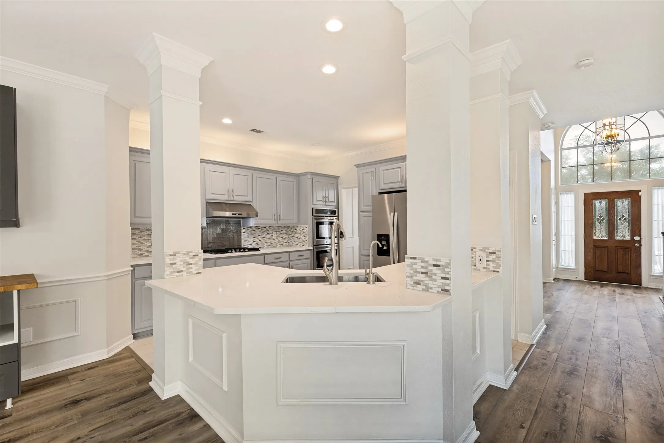 Open-concept kitchen featuring light gray cabinetry, built-in stainless steel appliances, a white countertop island with an undermount sink, and wood-finish flooring extending into the entry with a prominent arched window