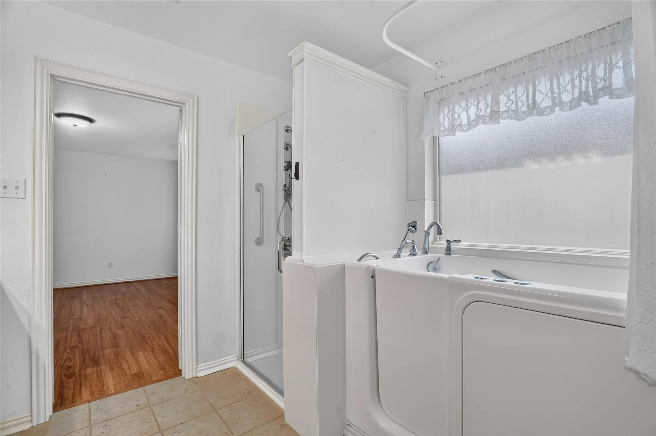 White walk-in tub with chrome fixtures, glass-enclosed shower, tile flooring, and a frosted window