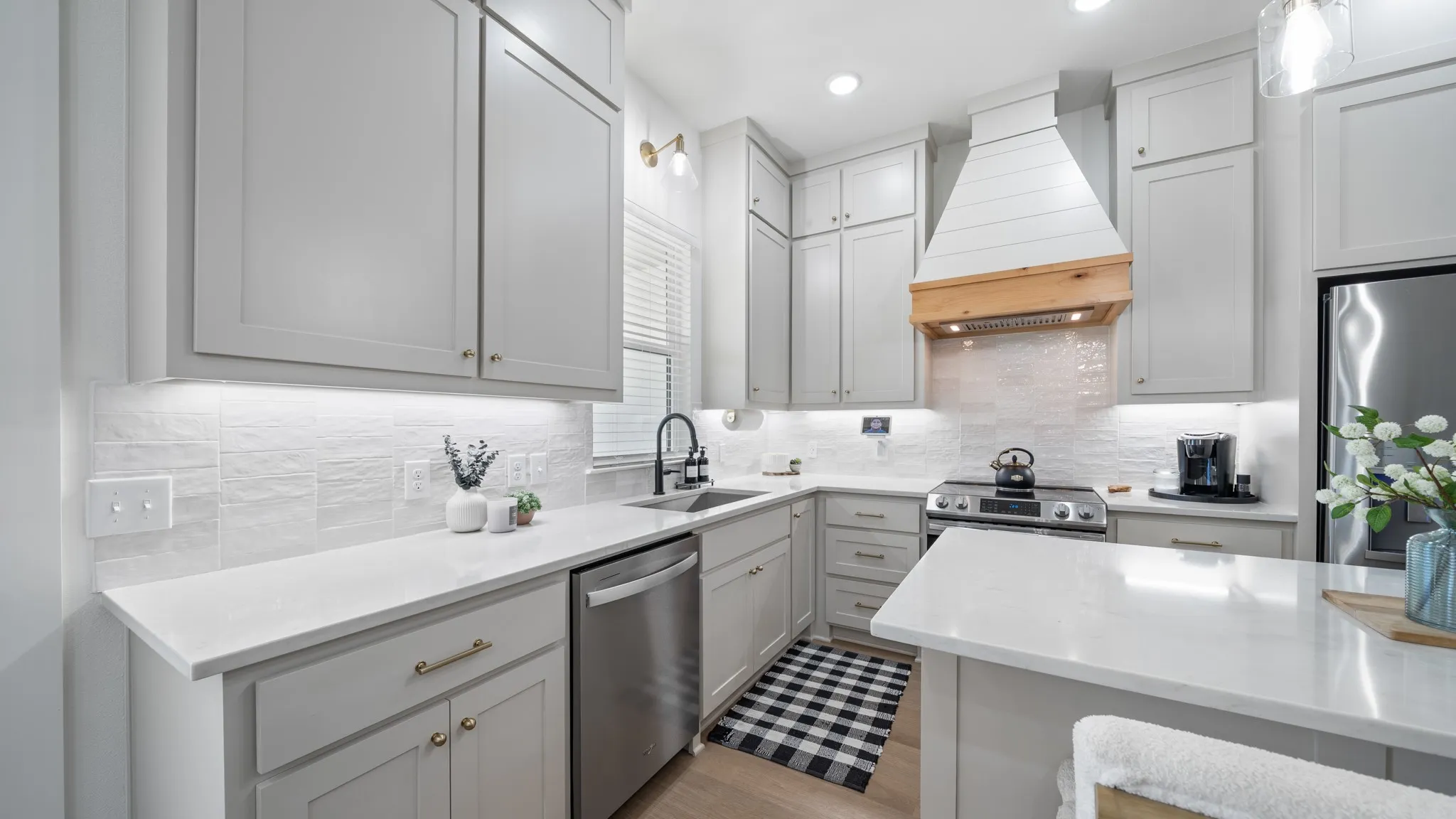 White paneled cabinetry with brass hardware, stainless steel appliances, a wood-finish range hood, and light-toned countertops