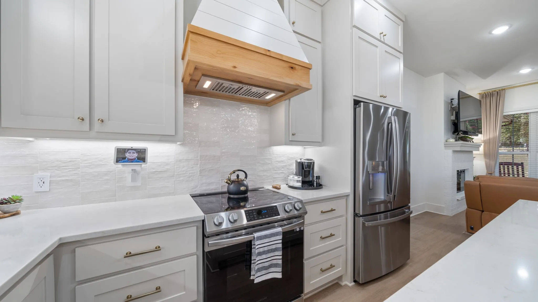 Modern kitchen featuring white cabinetry with brushed brass hardware, quartz countertops, stainless steel appliances including a French-door refrigerator and slide-in range, white subway tile backsplash, wood-finish hood surround, and wood-finish flooring that extends throughout the open-concept living area