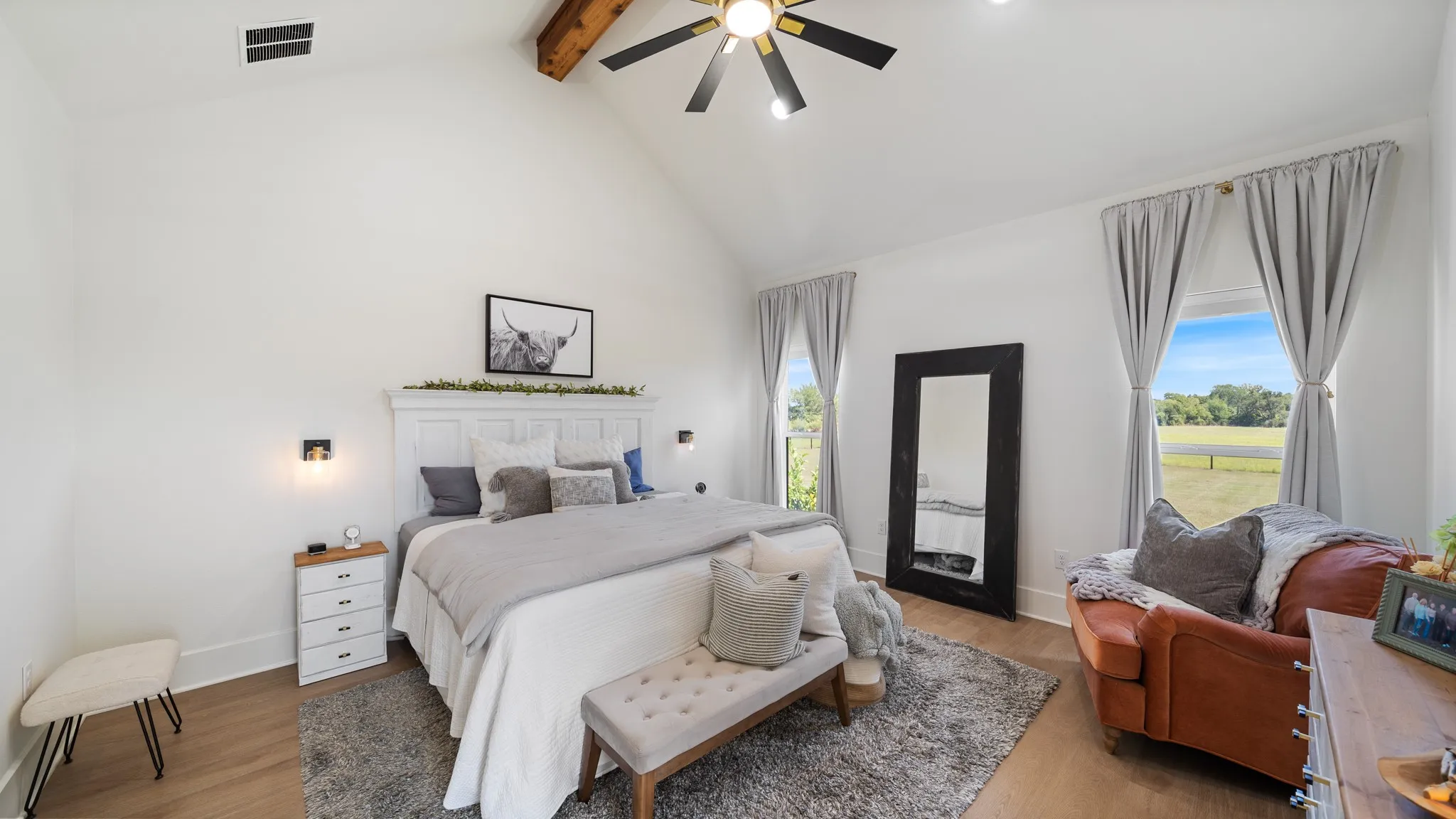 Vaulted ceiling with exposed wood beams, ceiling fan, wood-finish flooring, two large windows, and wall-mounted sconces