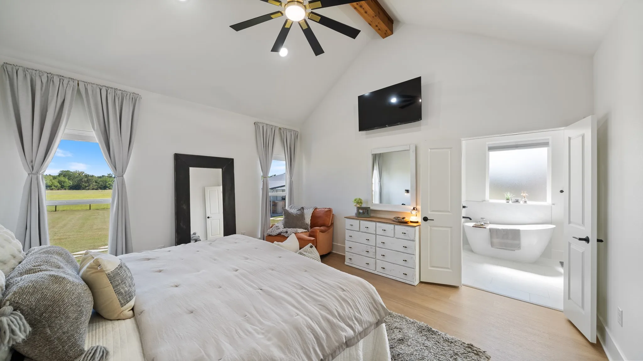 Vaulted ceiling with exposed wood beam, ceiling fan with light fixture, wood-finish flooring, white walls, and a freestanding soaking tub