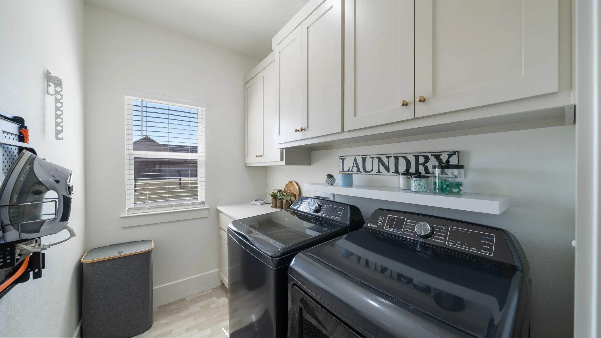 Dedicated laundry room featuring black front-load washer and dryer with modern digital controls, white shaker-style cabinetry with brushed nickel hardware, light-filled space with dual windows, decorative "LAUNDRY" signage, and neutral tile flooring