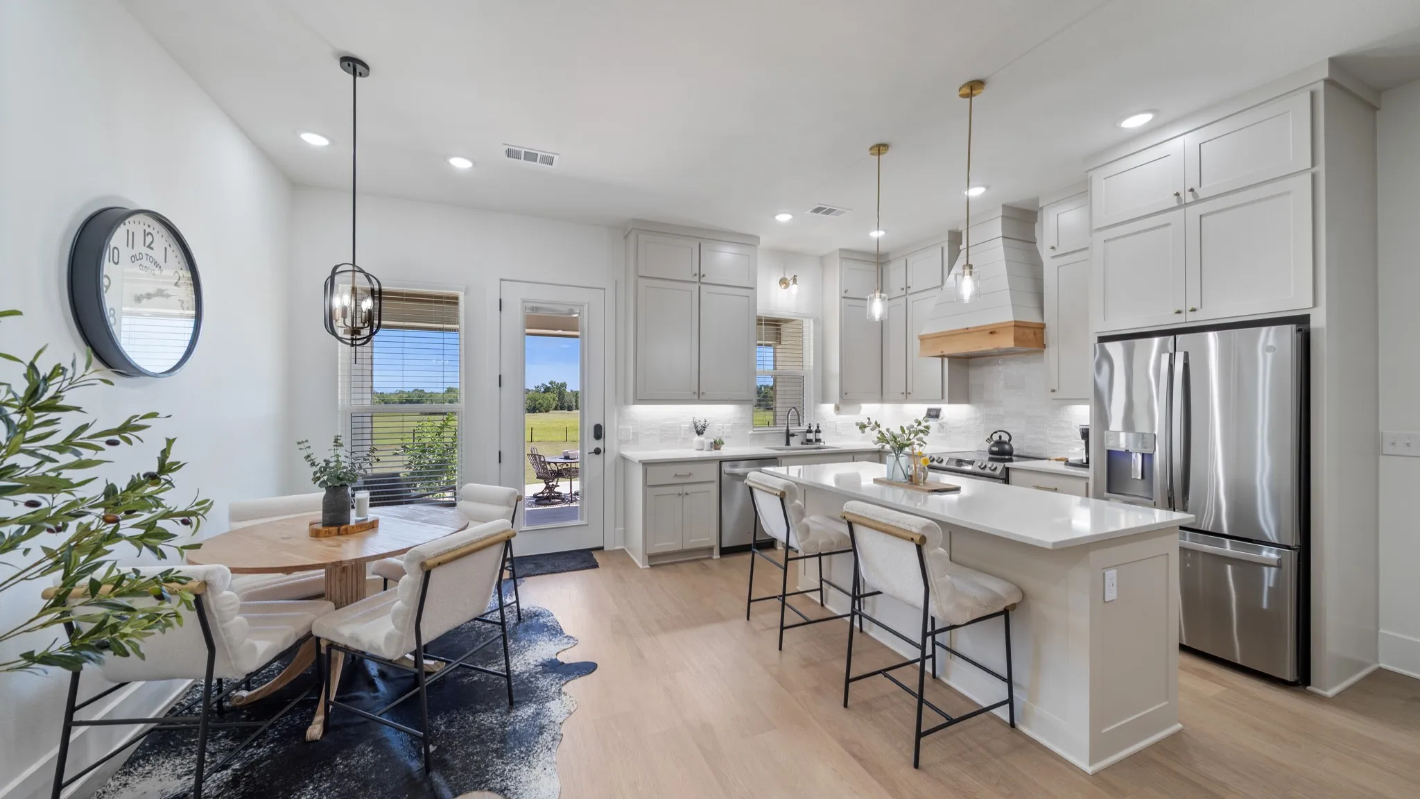 Open-concept kitchen and dining area featuring white cabinetry, stainless steel appliances, quartz countertops, a large center island with seating, pendant lighting, wood-finish flooring, and expansive windows with natural light and countryside views