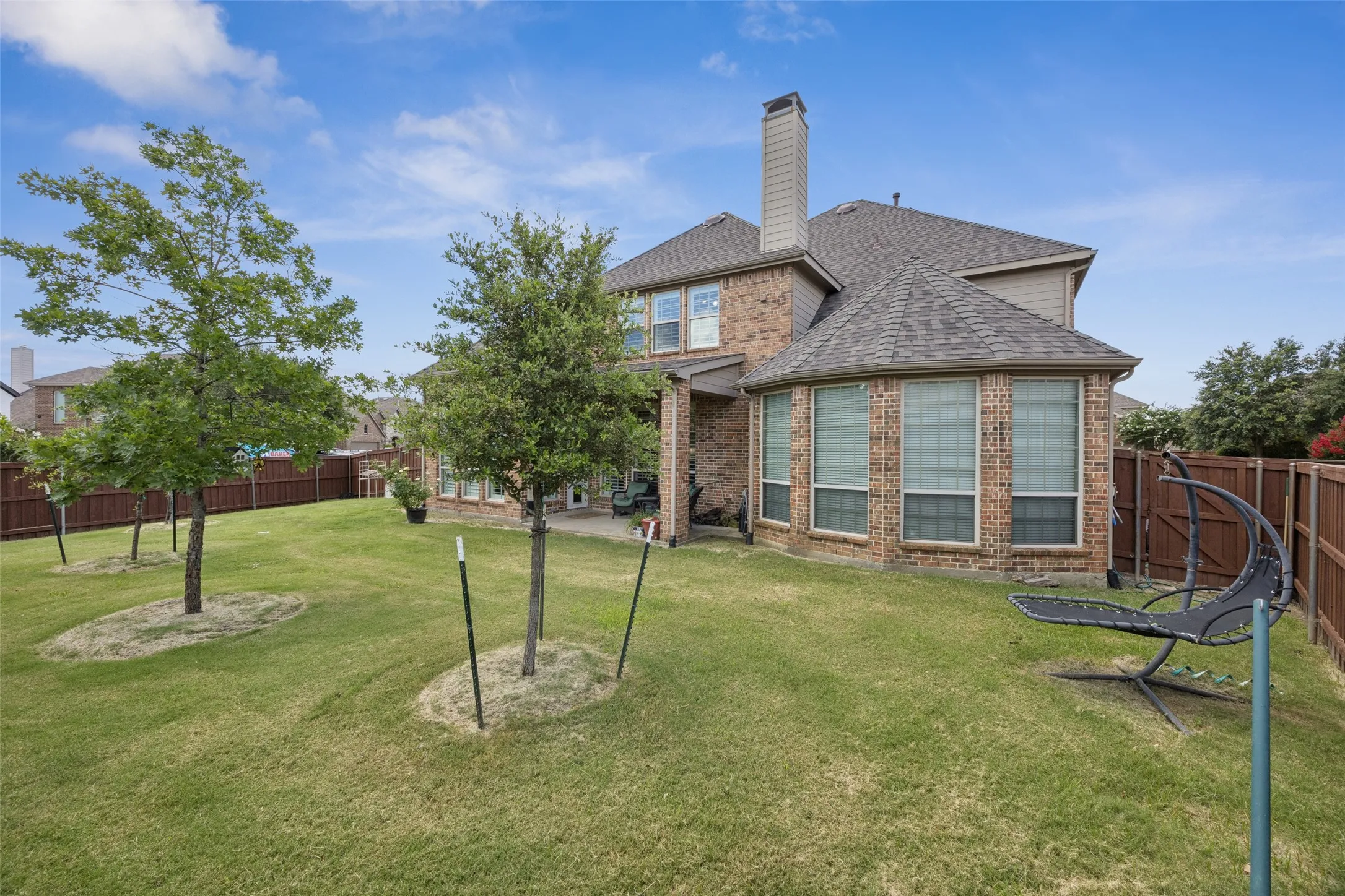 Expansive grassy yard with young trees, brick exterior, multiple windows, and a wood privacy fence