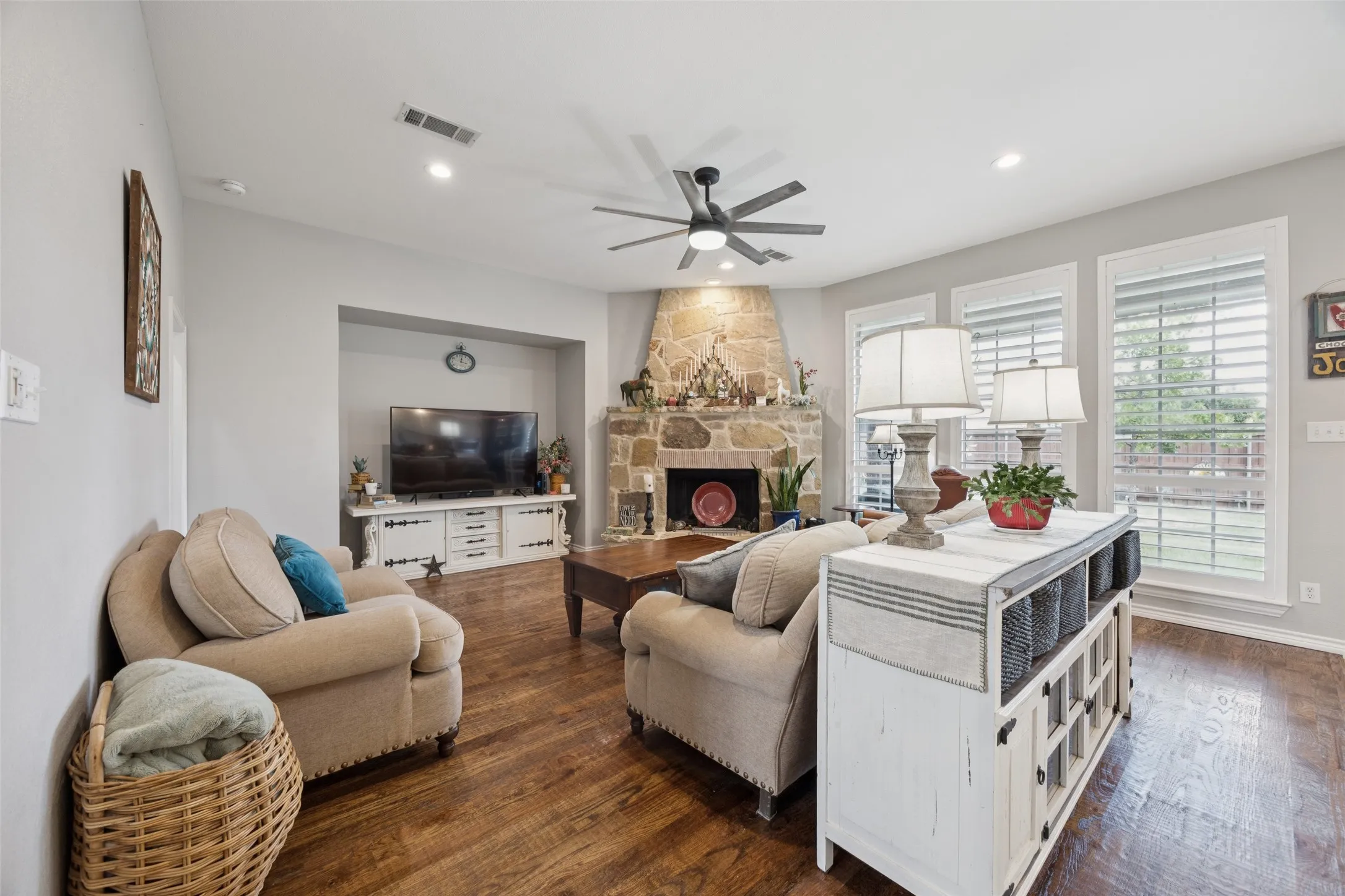 Stone-faced fireplace, wood-finish flooring, recessed lighting, ceiling fan, and multiple windows with plantation shutters