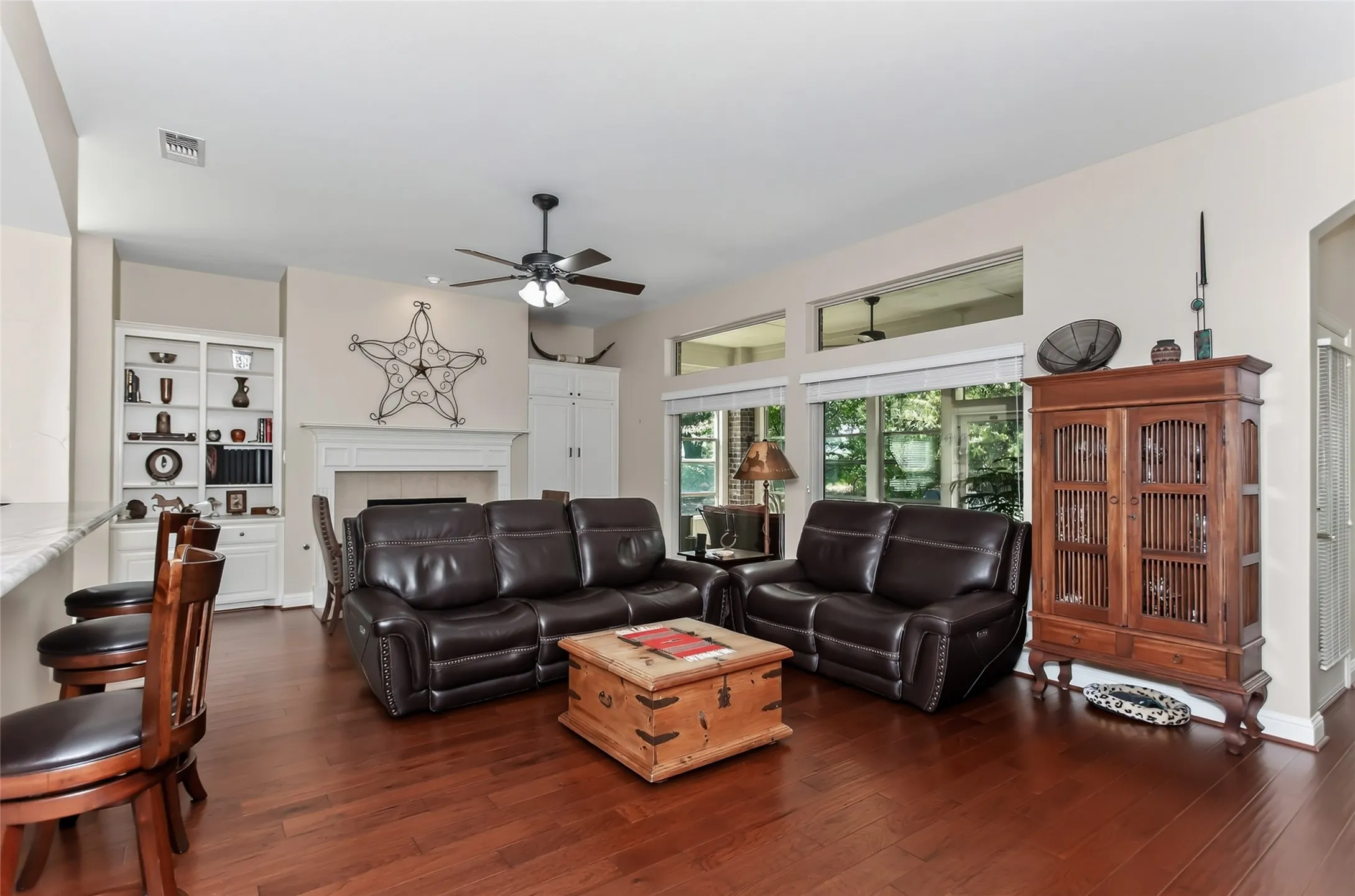 Wood-finish flooring throughout, featuring a fireplace with a mantel, built-in shelving, a ceiling fan, and large windows with transom details