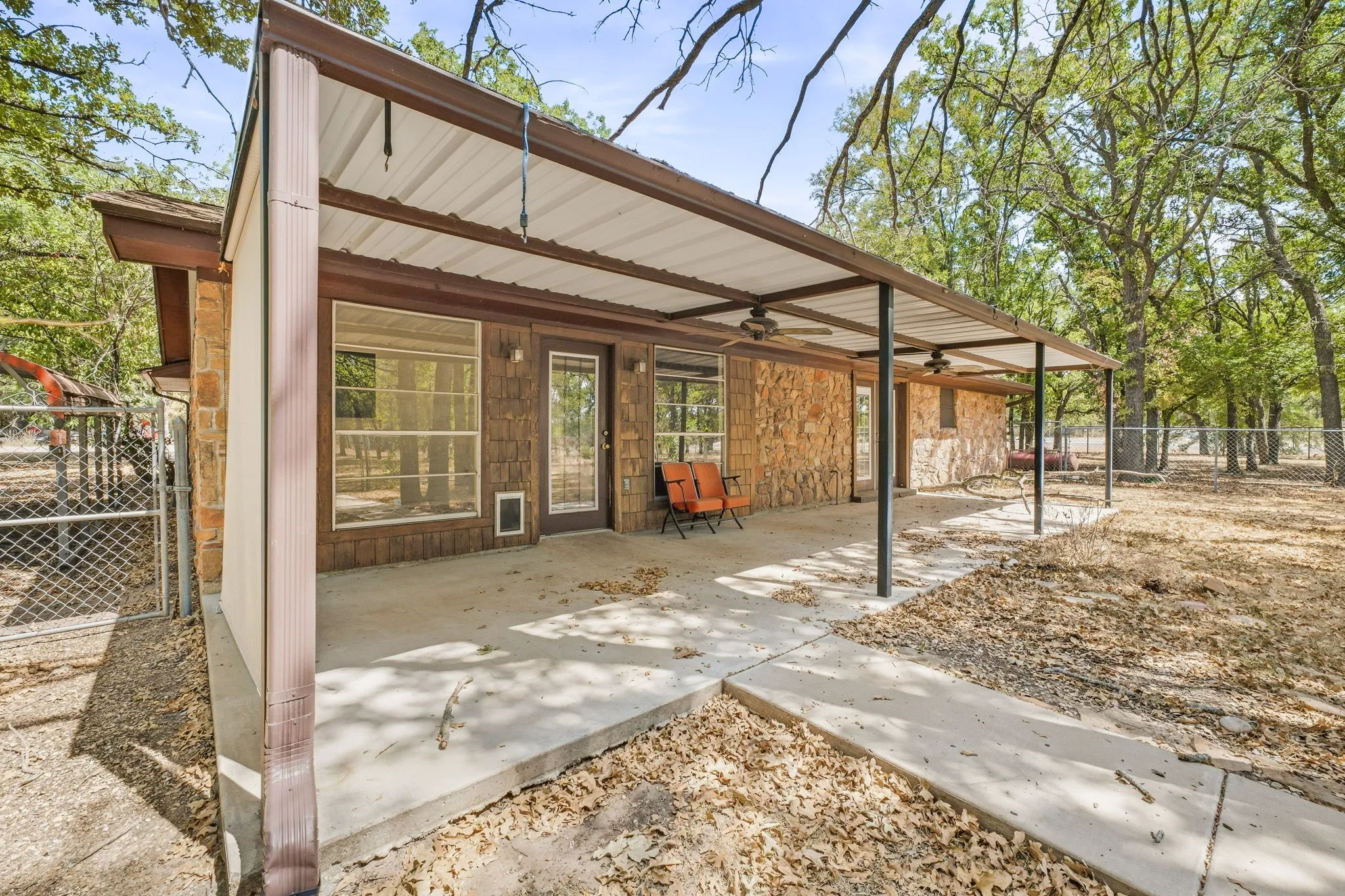 Expansive concrete patio featuring a metal awning with ceiling fans, a stone exterior, and a fenced perimeter