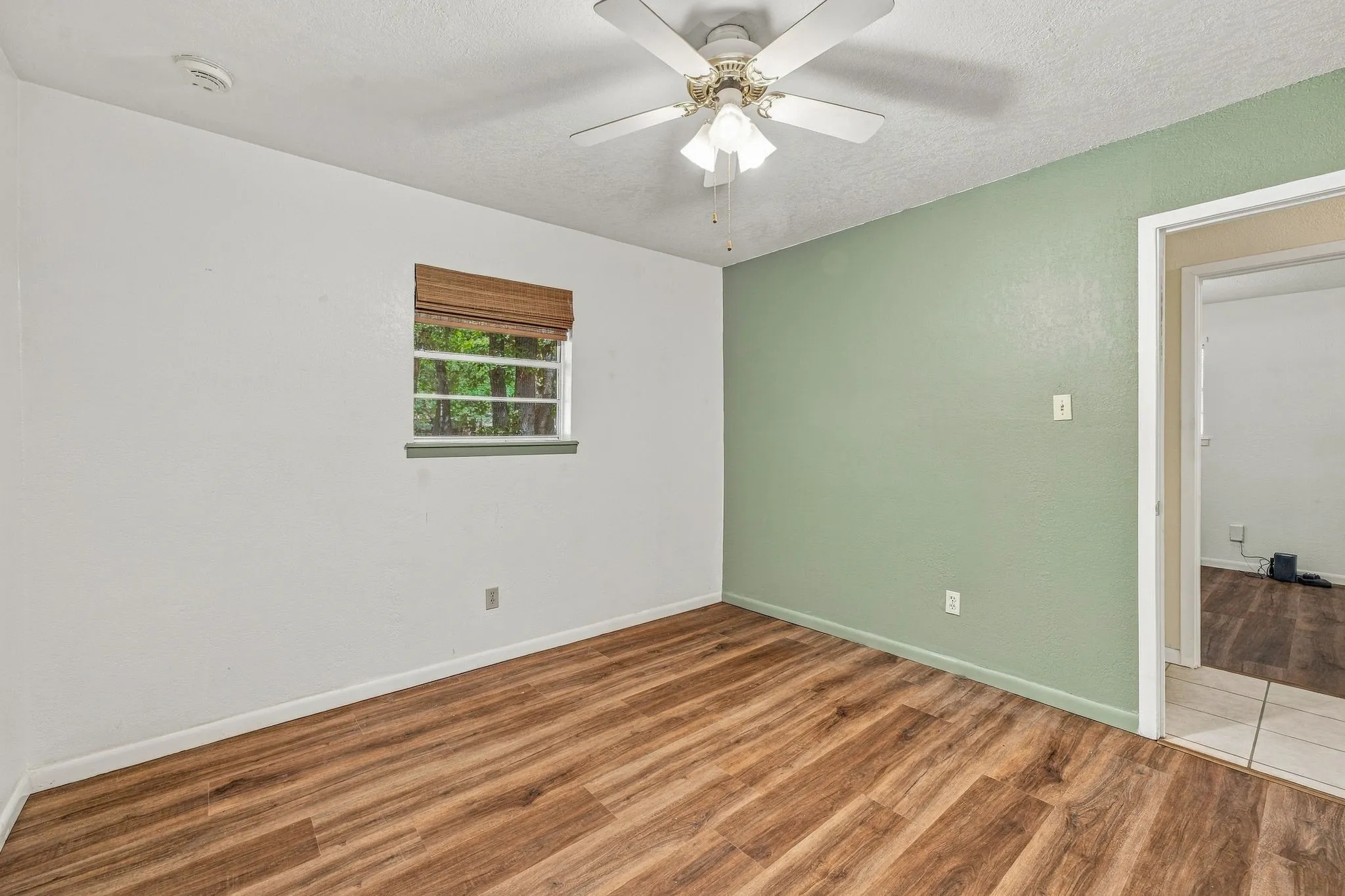 Wood-finish flooring, ceiling fan with integrated lighting, natural light window with blinds, and contrasting accent wall