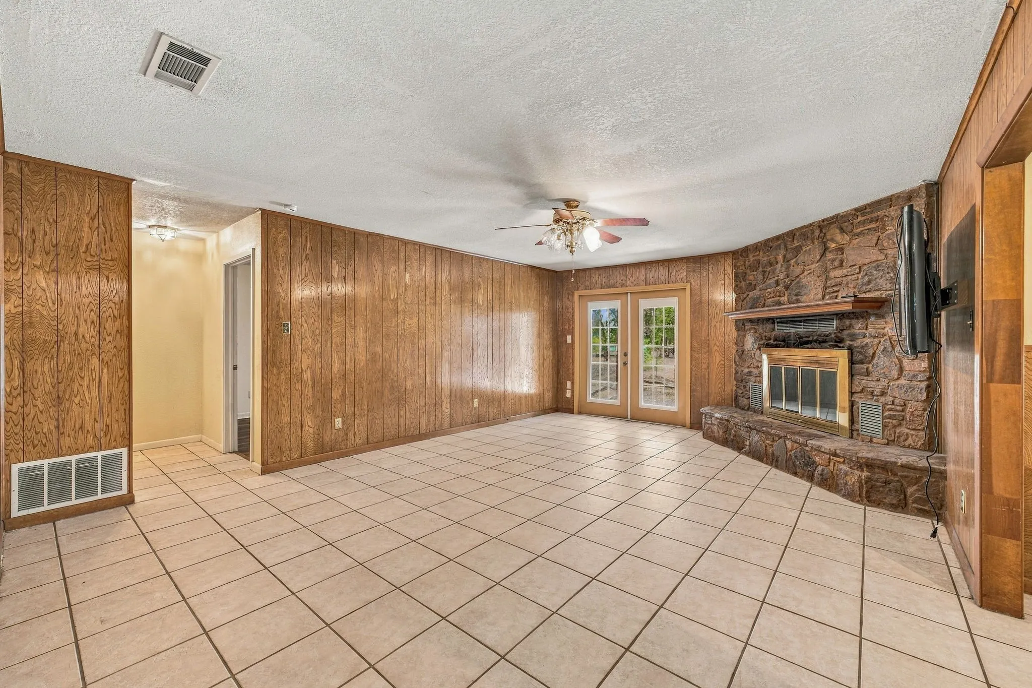 Tiled flooring throughout, wood paneling, a stone-clad fireplace with hearth, a ceiling fan, and French doors
