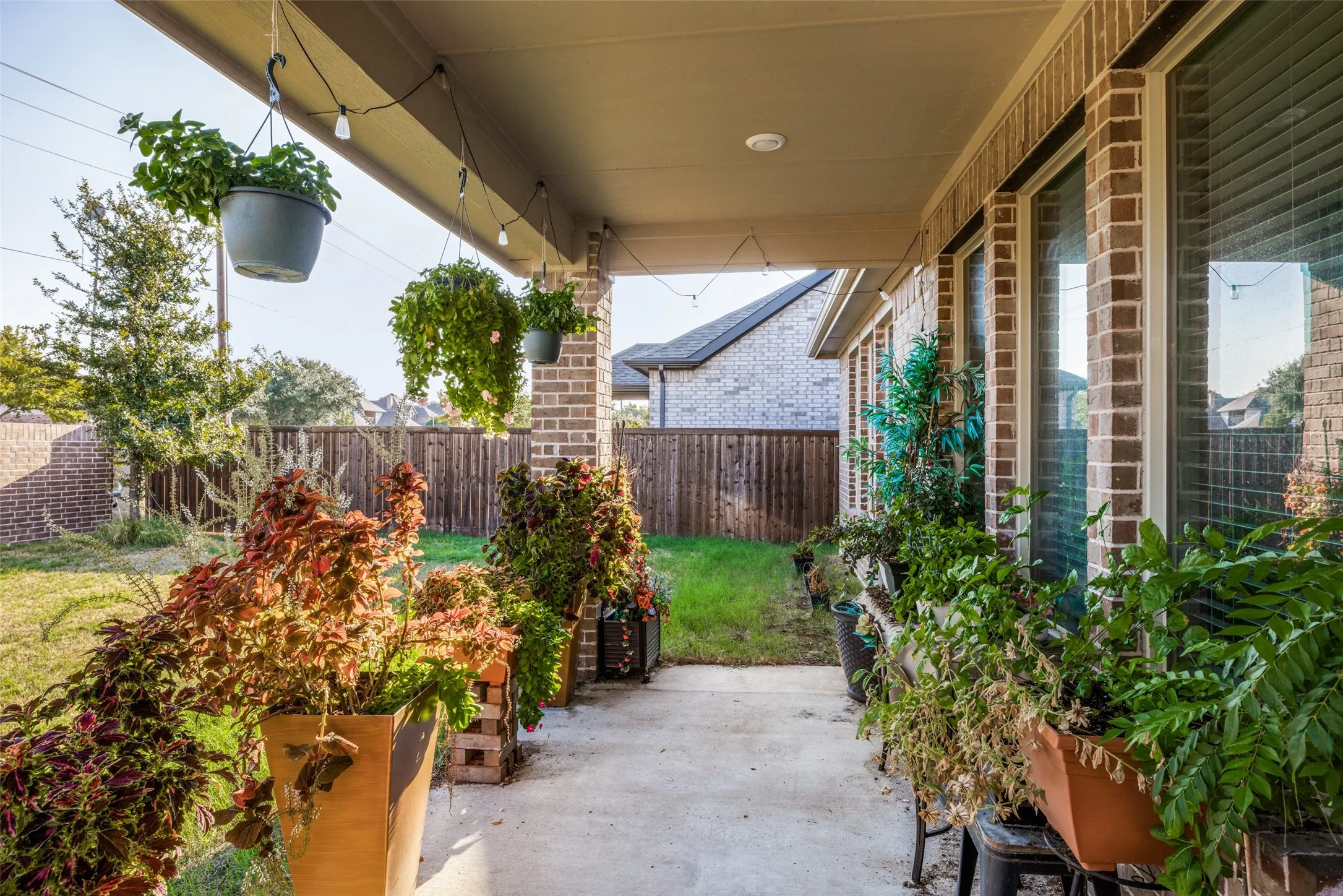 Covered patio with string lighting, brick exterior wall, multiple large windows, concrete patio surface, and wood privacy fencing