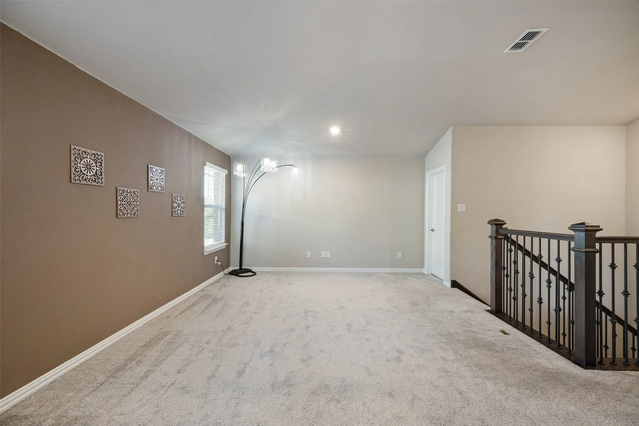Expansive carpeted room featuring a window with blinds, recessed ceiling lighting, and a dark wood and iron railing