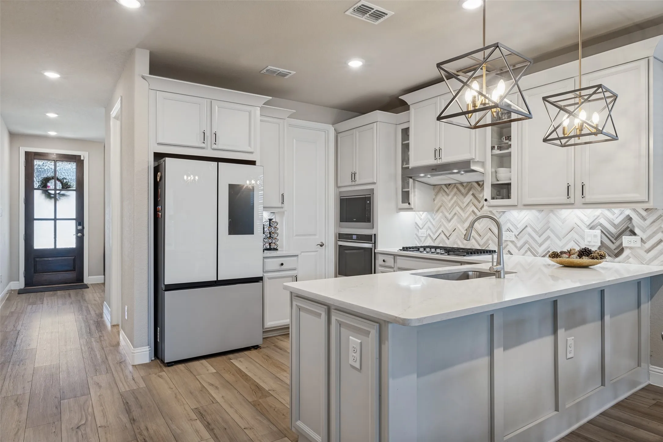 Kitchen featuring wood-finish flooring, white cabinetry, a large center island with a light-colored countertop, built-in appliances, and geometric pendant lighting