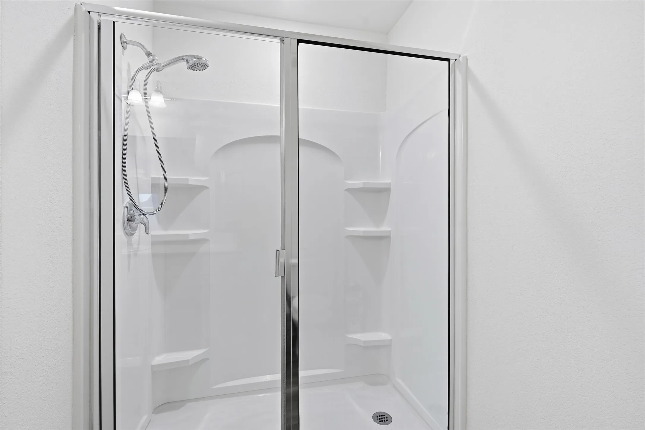 White shower stall with sliding glass door, integrated shelving, and a handheld showerhead