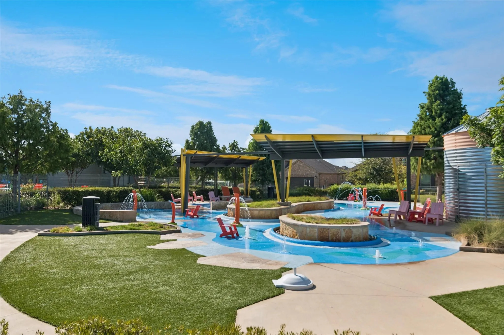 Splash pad featuring multiple water features, integrated seating, and shade structures, surrounded by artificial turf and mature landscaping