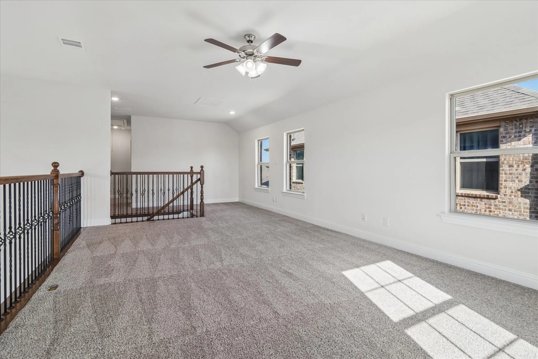 Expansive carpeted loft area with wrought iron and wood balustrade, multiple windows, and a ceiling fan with integrated lighting
