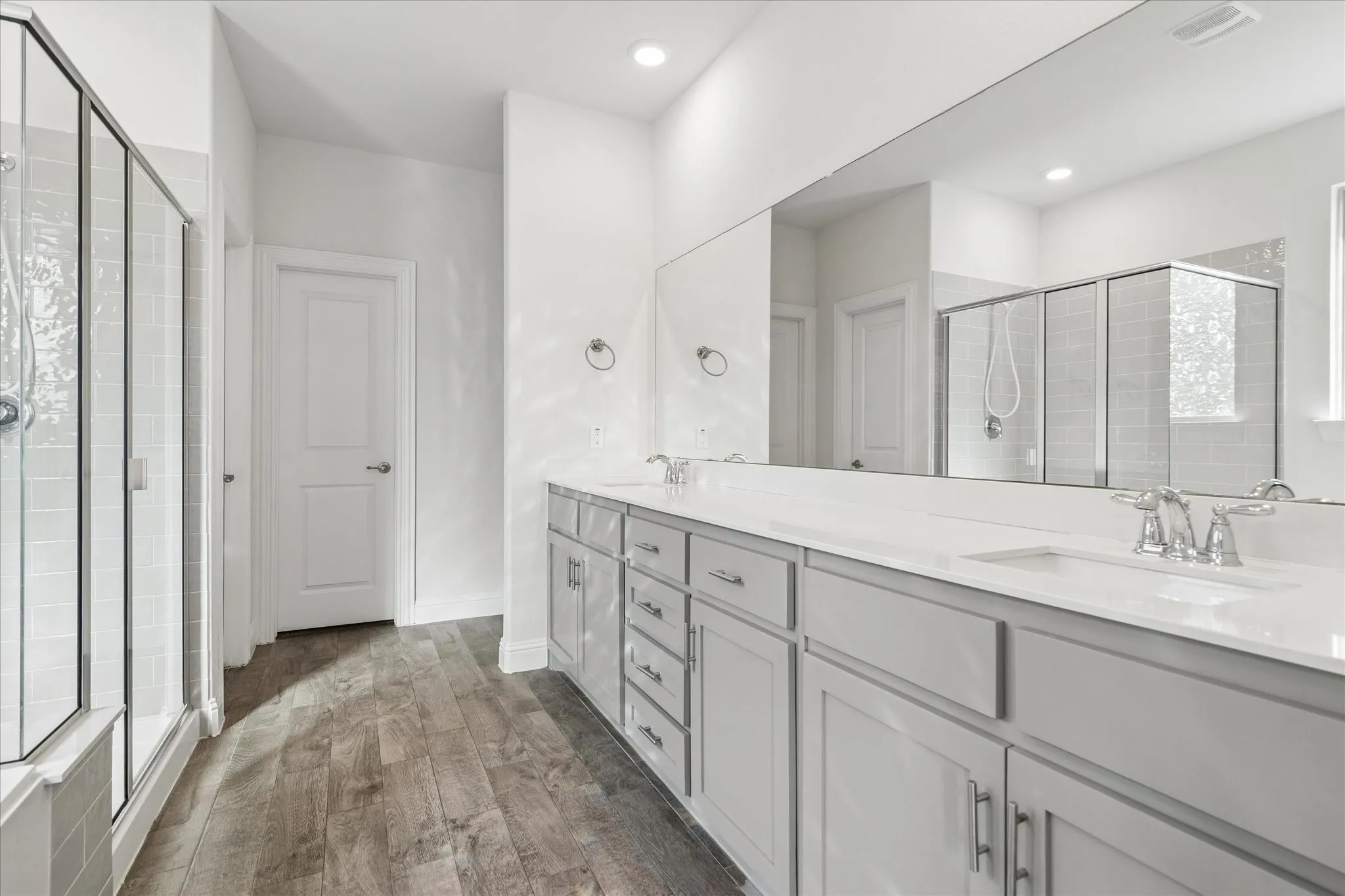 Double vanity with white countertops, wood-finish flooring, glass-enclosed shower, and a large wall mirror
