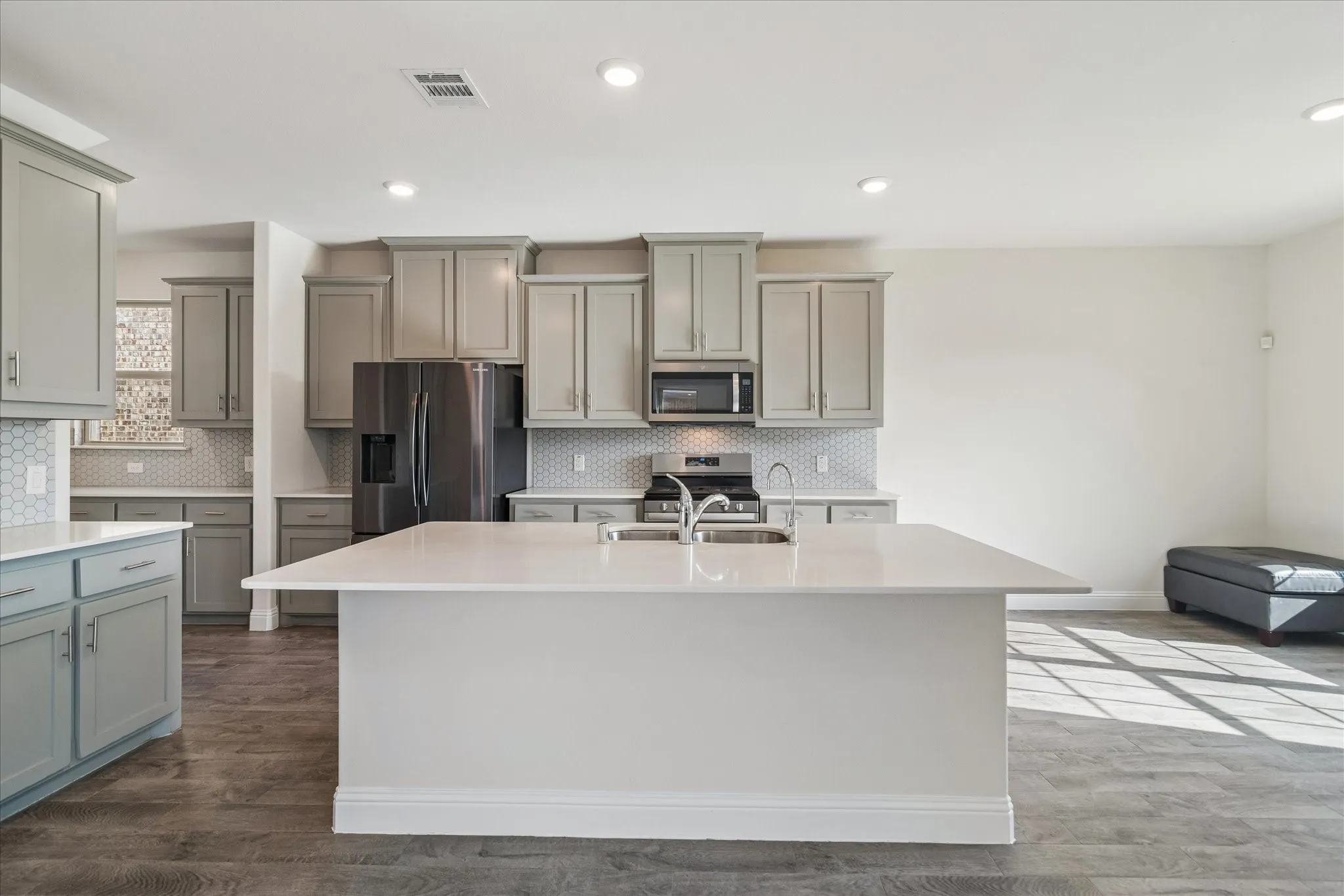 Kitchen featuring an expansive island with a stainless steel double basin sink, light-toned countertops, gray cabinetry, and hexagonal tile backsplash