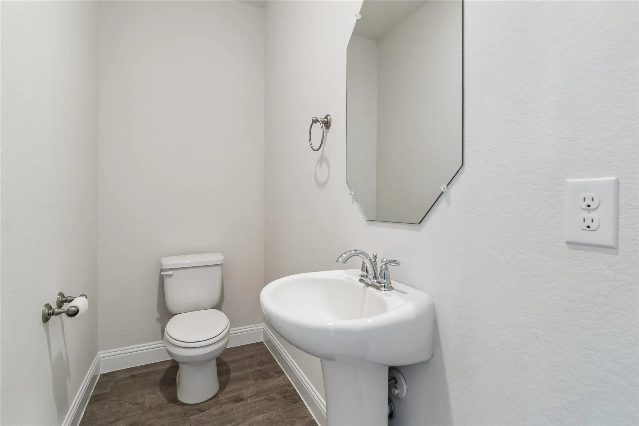 Pedestal sink with chrome fixtures, coordinating toilet, wood-finish flooring, and a decorative wall mirror