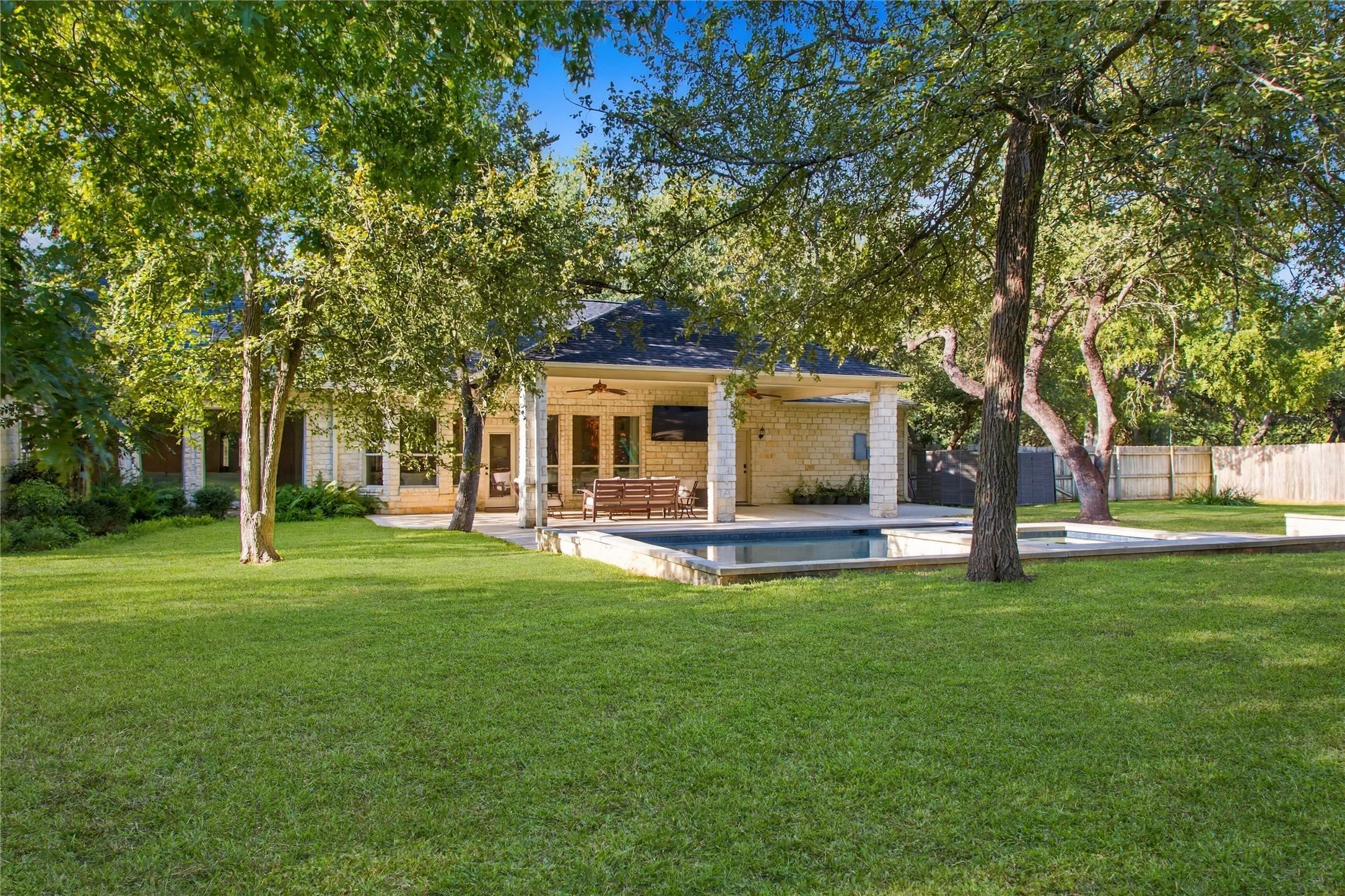 Expansive green lawn with mature trees, stone exterior, covered patio, and in-ground swimming pool