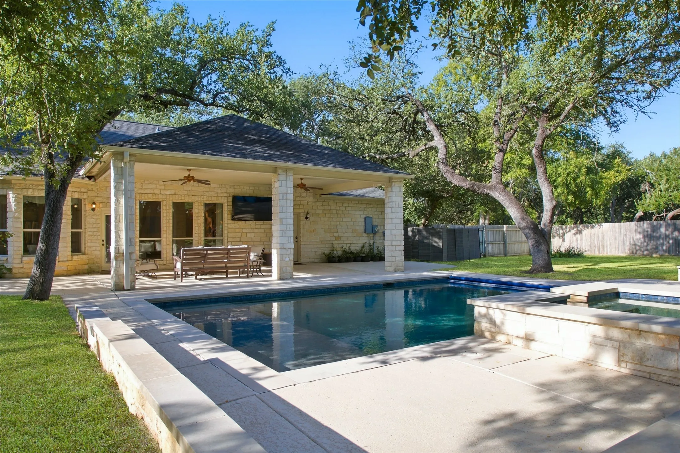 Outdoor pool with adjacent spa, covered patio with stone columns, and stone exterior