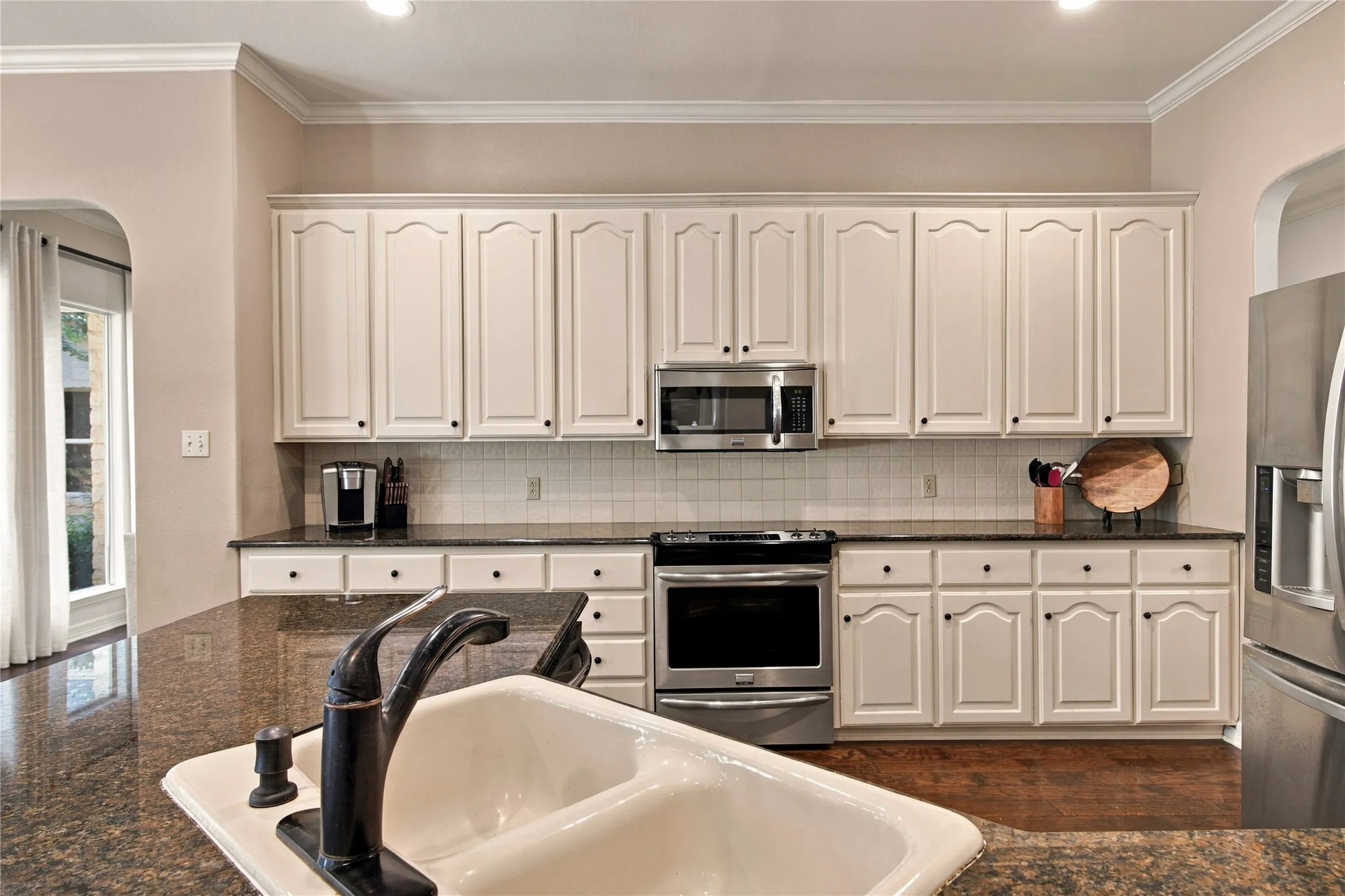 White cabinetry with black hardware, stainless steel appliances, dark stone countertops, tile backsplash, and wood-finish flooring