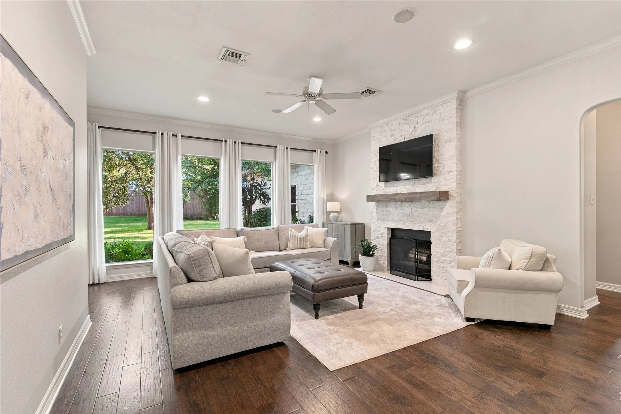 Expansive living area featuring rich wood-finish flooring, a white brick-clad fireplace with a substantial wood mantel, a ceiling fan, recessed lighting, and multiple windows overlooking greenery
